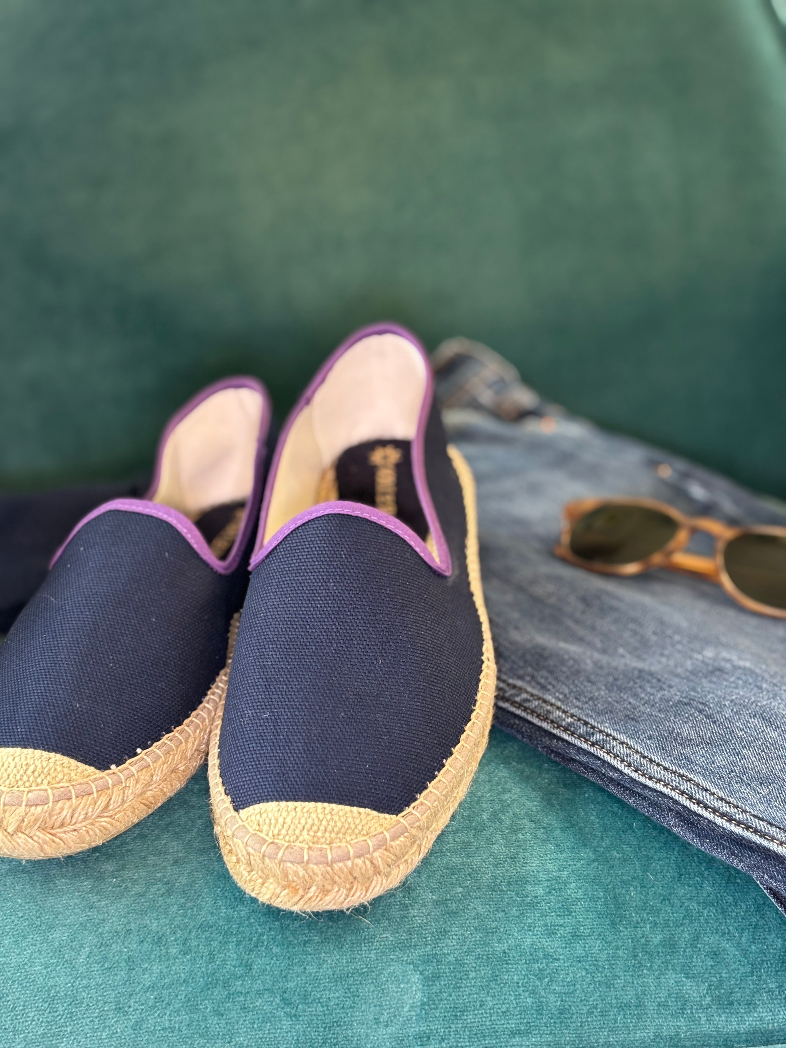 Mens blue espadrille loafers beside a pair of brown sunglasses and denim trousers on a green background