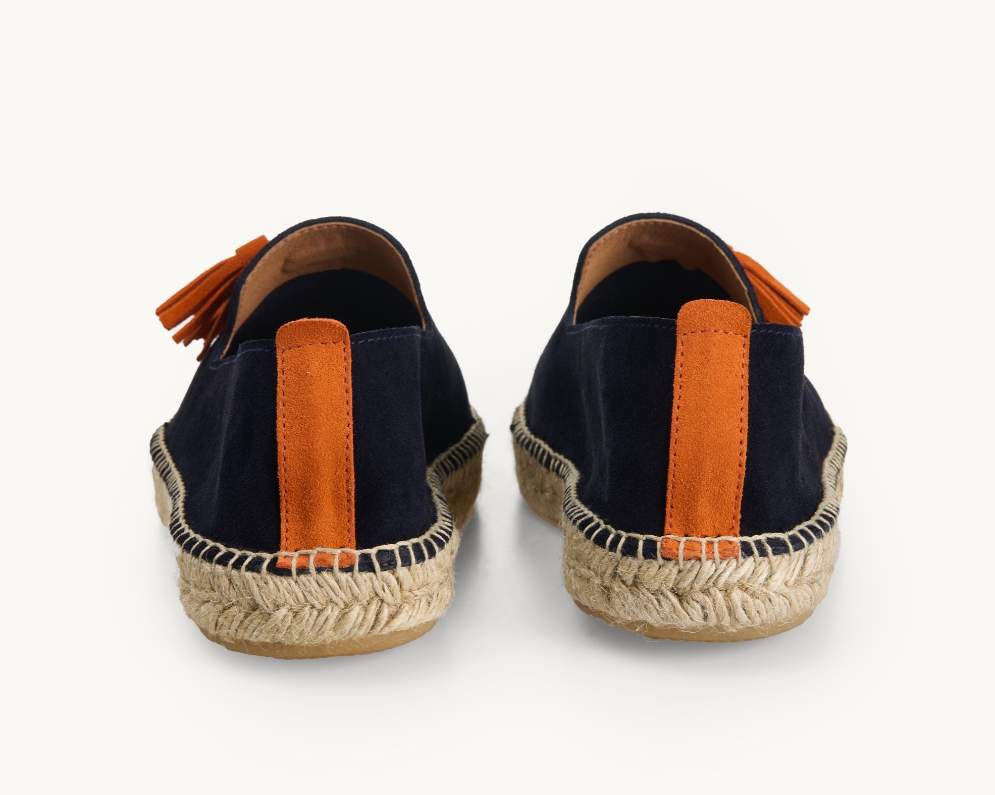 A pair of navy suede men's espadrille loafers. Only the back details of the heels are shown. A stripe of orange suede about a centimetre wide runs straight up the back heel. Stitching around the edge of the upper to join it to the traditional jute sole. The side of the orange tassels are shown.
