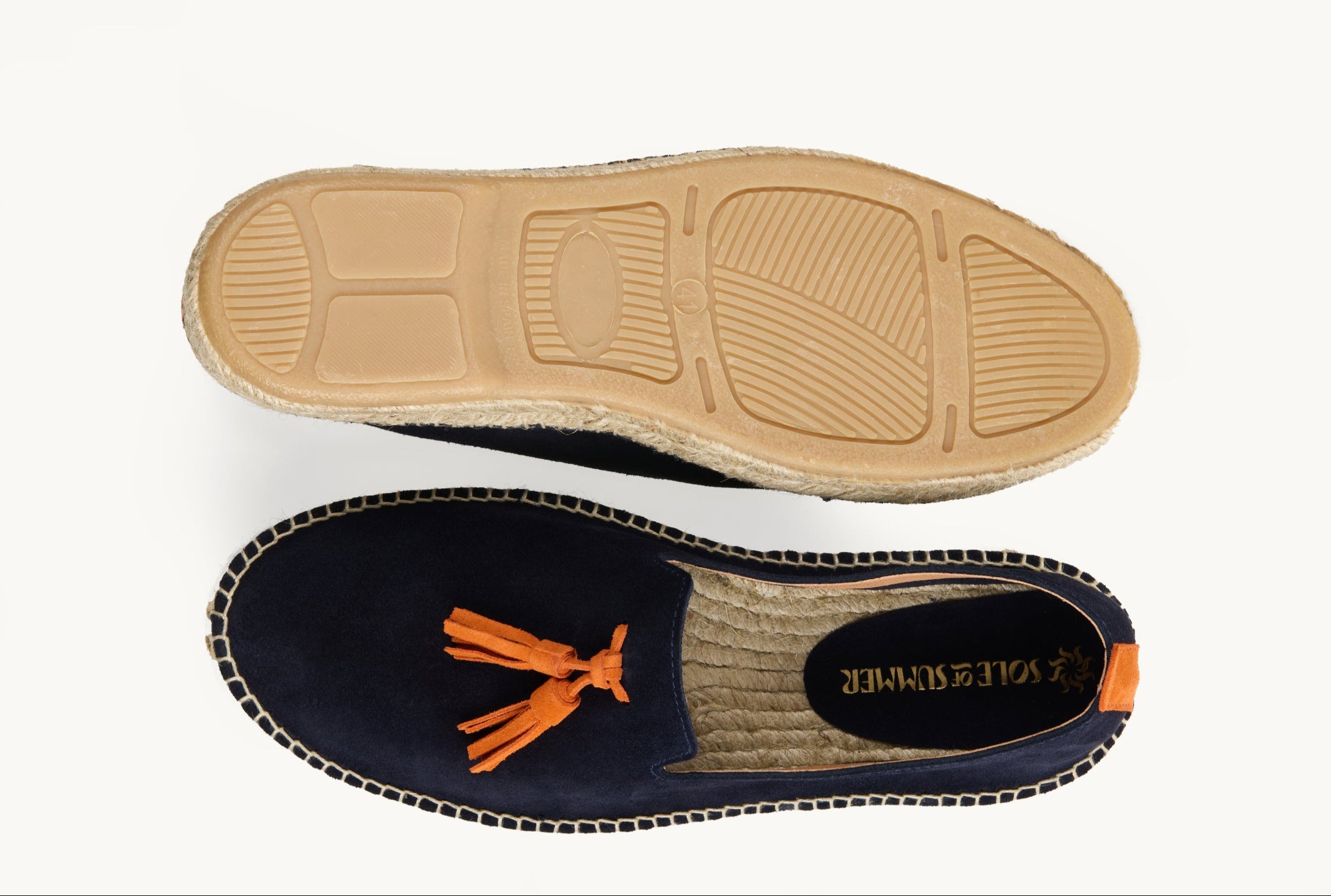 A pair of men's navy blue suede loafer espadrilles. One of the footwear is turned over to show the underneath. It has a full rubber covering and the other shoe shows the inner, unlined jute insole and the Sole of Summer logo printed on a small piece of navy blue suede. The other shoe is the right way up and shows the orange suede tassels on the front of the footwear and a orange suede stripe up the heel.