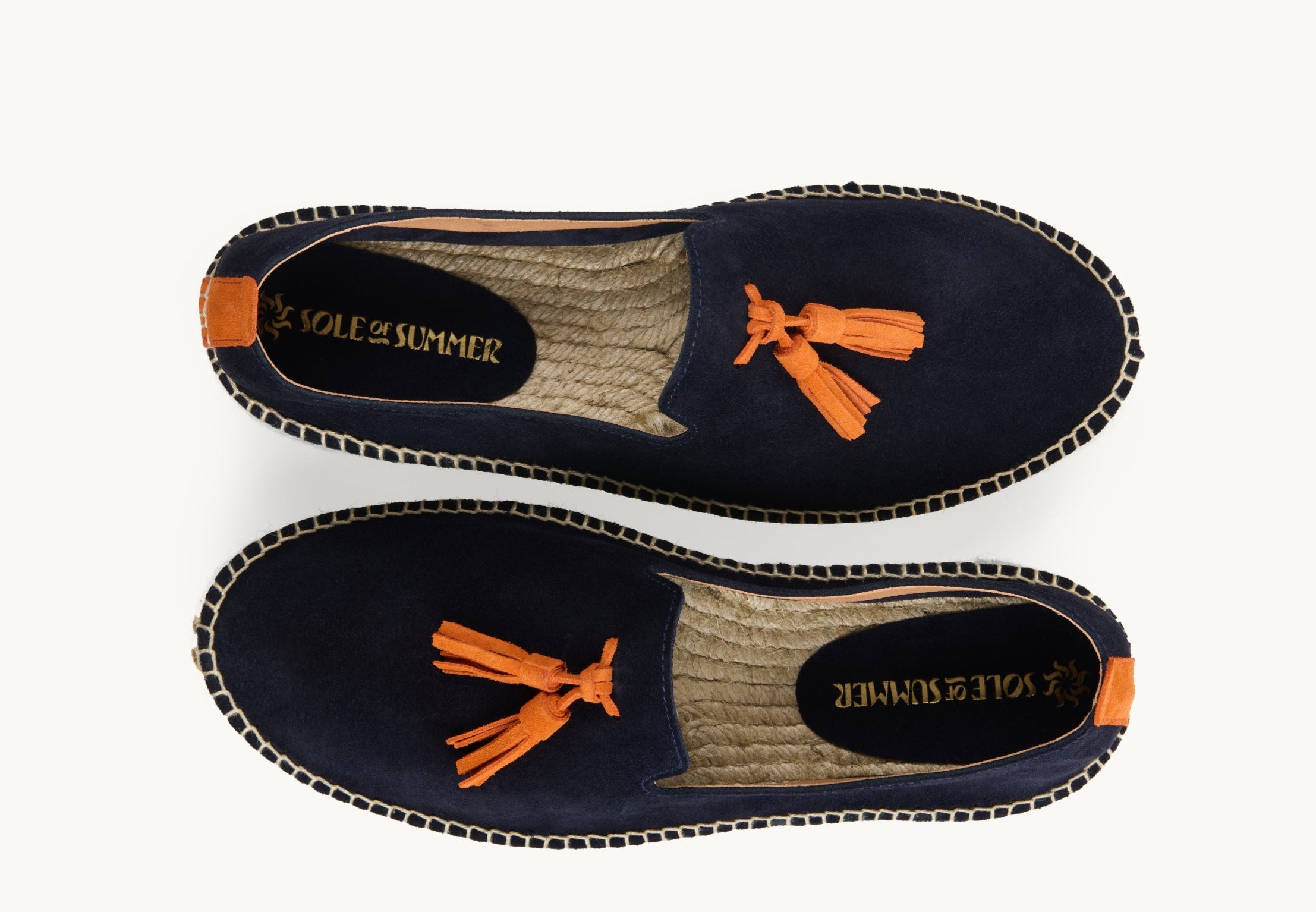A pair of men's loafer espadrilles. Arial view. Navy blue suede upper with orange suede tassels on the front. Insole is unlined, jute inside with a small piece of orange suede printed with Sole of Summer logo in gold. Stripe of orange suede about 1cm wide up the heels.