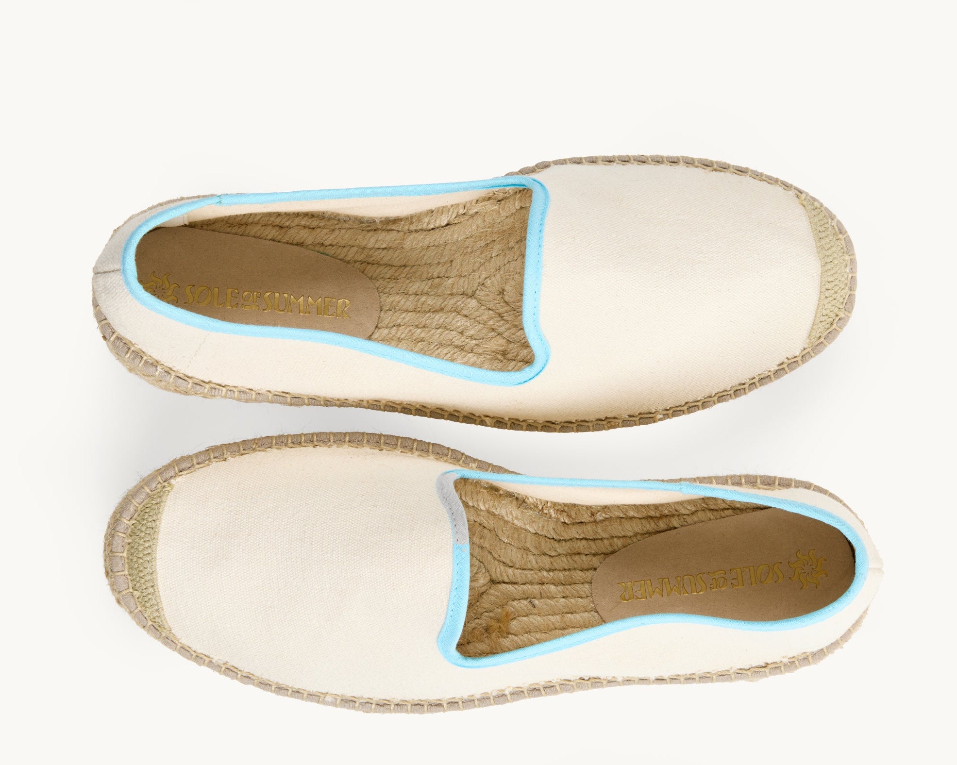 A pair of men's white cotton loafer espadrilles, the opening of the shoe is edged in a narrow light blue ribbon. One of the footwear is turned over to show the underneath. It has a full rubber covering and the other shoe shows the inner, unlined jute insole and the Sole of Summer logo printed on a small piece of cream suede.