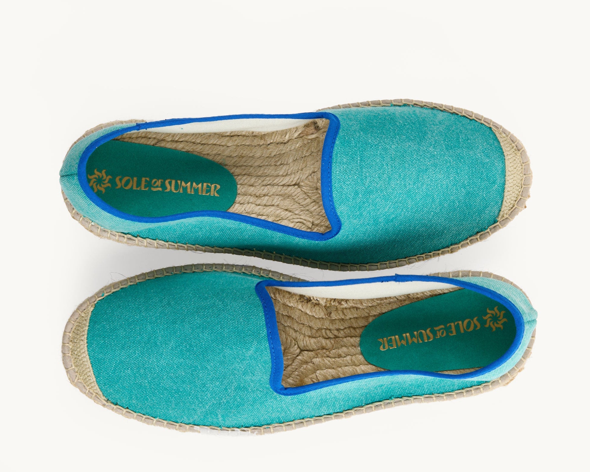A pair of men's green cotton loafer espadrilles, the opening of the shoe is edged in a narrow royal blue ribbon. One of the footwear is turned over to show the underneath. It has a full rubber covering and the other shoe shows the inner, unlined jute insole and the Sole of Summer logo printed on a small piece of green suede.