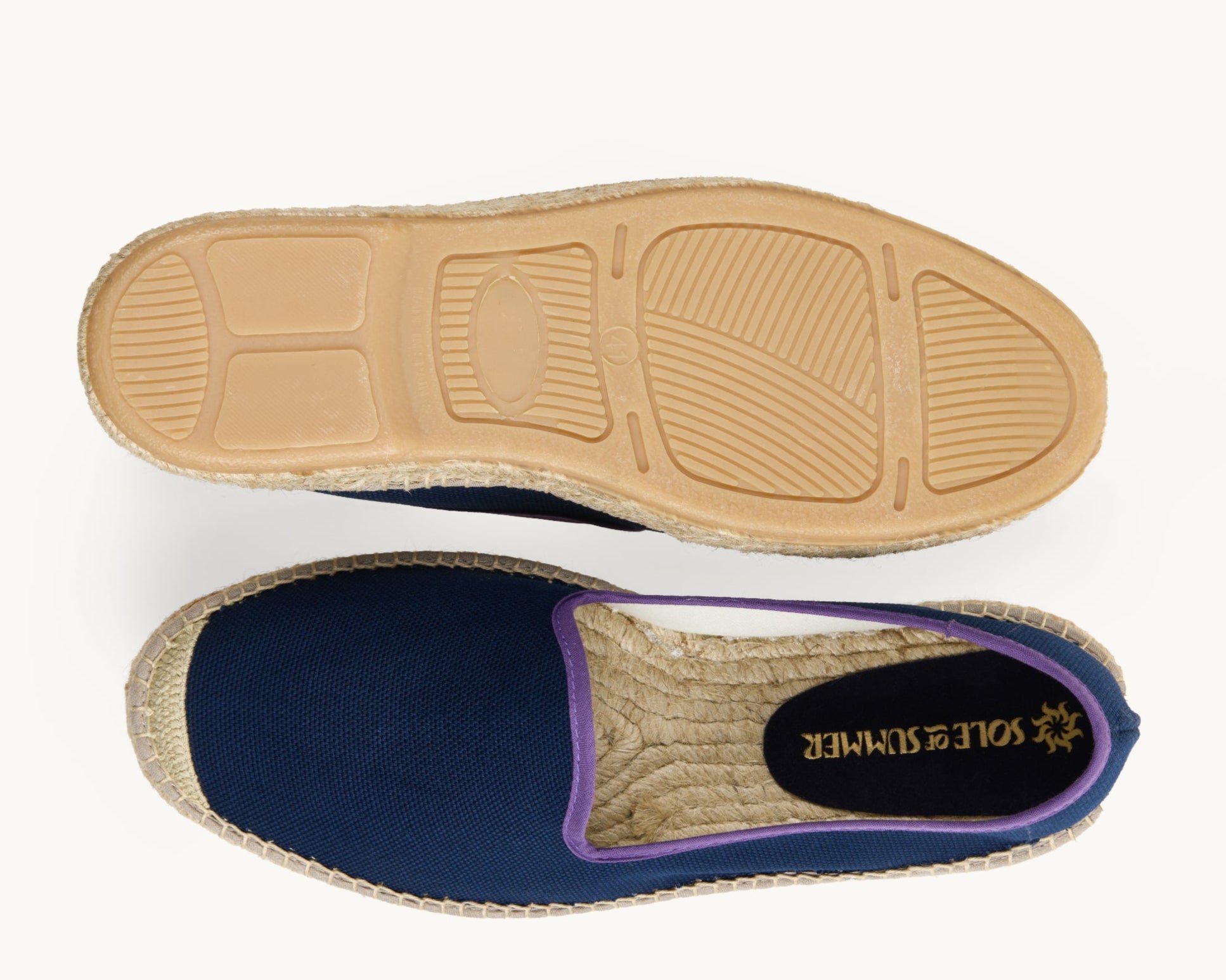 A pair of men's navy cotton loafer espadrilles, the opening of the shoe is edged in a narrow purple ribbon. One of the footwear is turned over to show the underneath. It has a full rubber covering and the other shoe shows the inner, unlined jute insole and the Sole of Summer logo printed on a small piece of blue suede.