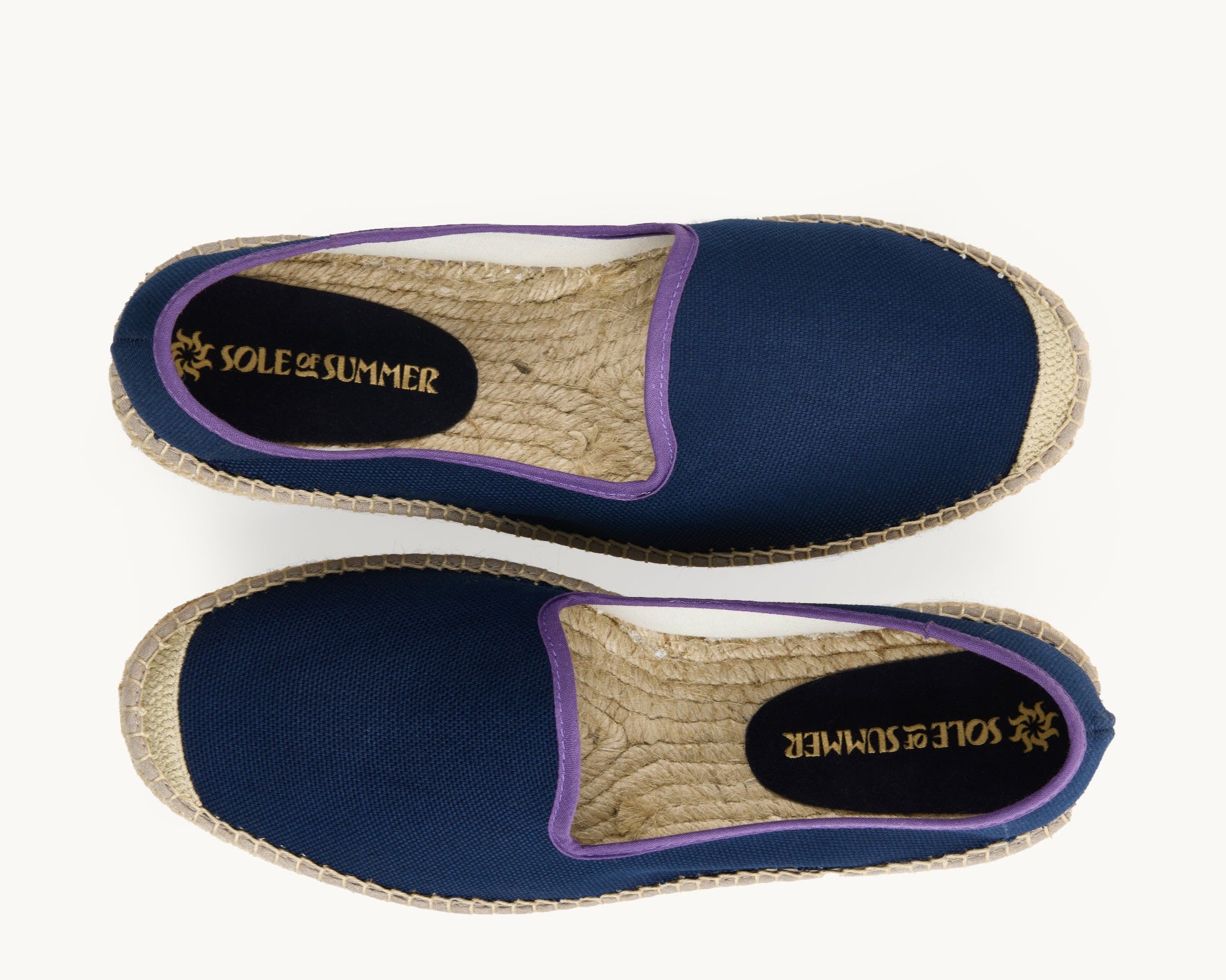 A pair of men's loafer espadrilles. Arial view. Navy blue cotton upper with narrow purple trim around the opening of the shoe. Insole is unlined, jute inside with a small piece of navy suede printed with Sole of Summer logo in gold.