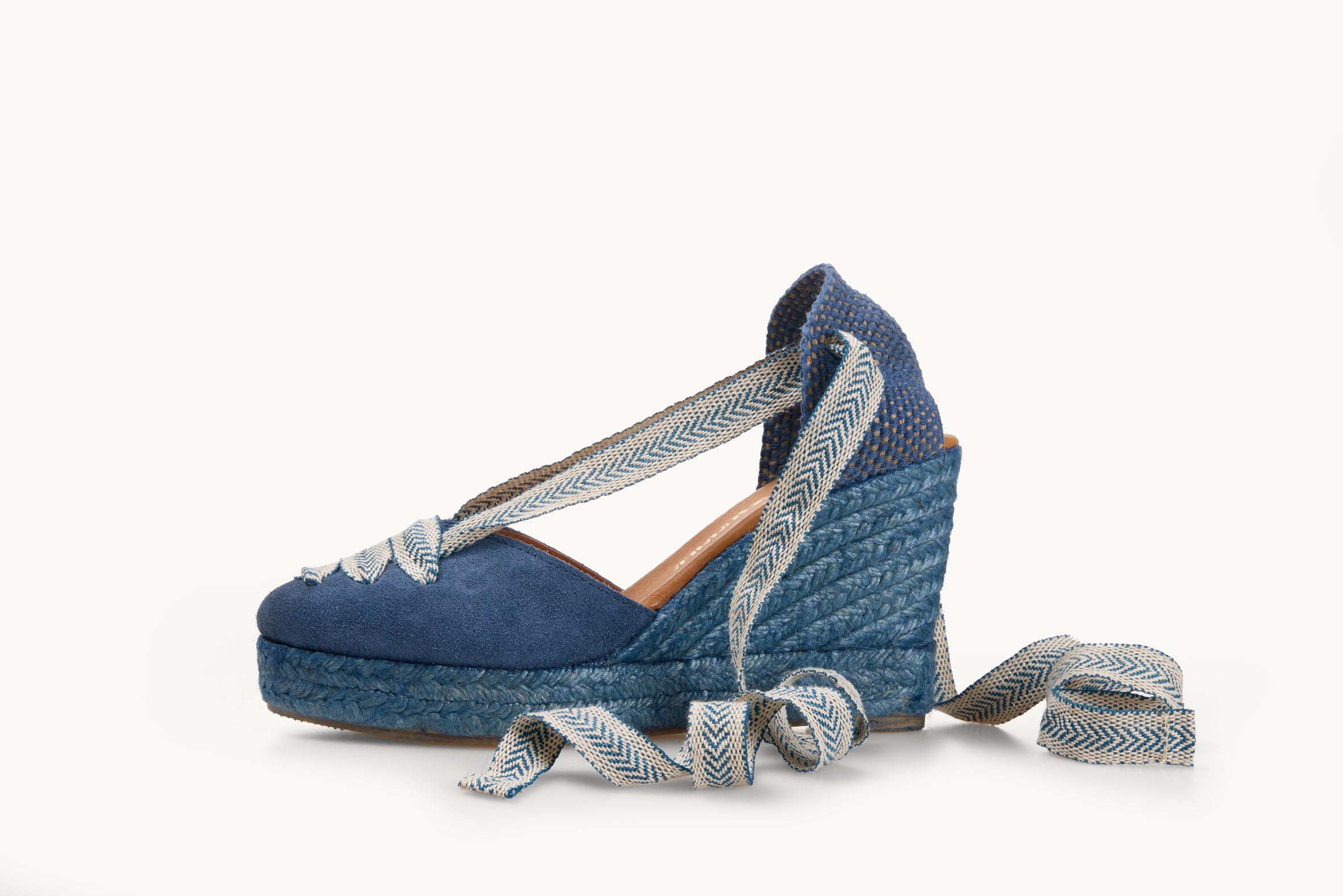 One iris denim blue suede wedge espadrille shoe, side view. It has a matching blue jute sole. Across the almond shaped toe is three rows of blue ribbon with white arrow head pattern. The ankle ties are in the same ribbon material and are untied and loose to the side and a ribbon joining the toe to the heel.