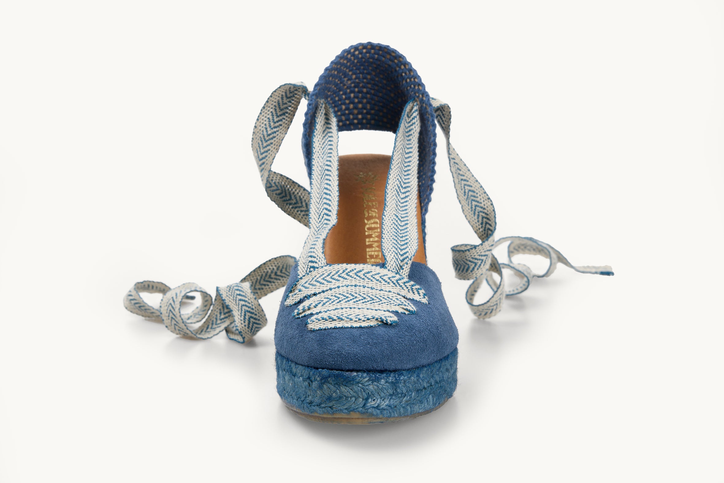 One iris denim blue suede wedge espadrille displayed with matching blue and white arrow head pattern ribbons at the heel, left loose and untied on either side. The base is made of matching blue Jute. The ribbons are also in three lines across the almond shaped toe. Two ribbons go from the toe to the heel on the left and right. The insole is shown in tan leather with the Sole of Summer logo printed in gold.