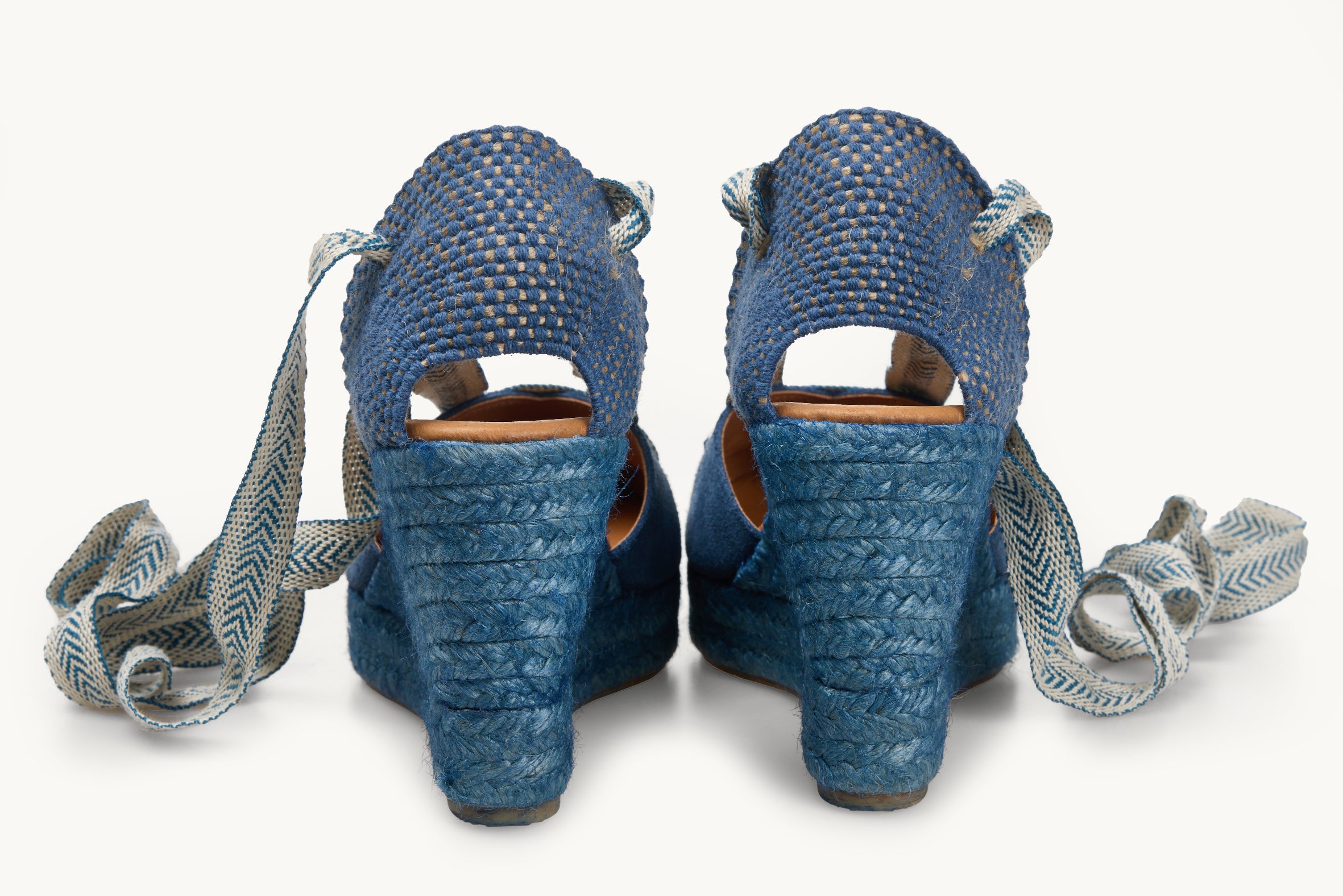 A pair of iris denim blue wedge espadrilles looking at the heel details and the blue jute soles are visible with the blue and white arrow head pattern ribbons untied loose next to the shoes.