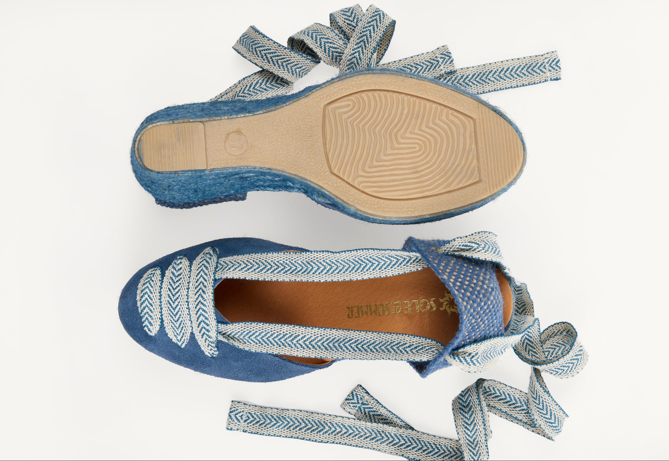 A pair of women's iris denim blue suede wedge espadrilles with blue and white arrow head pattern ribbons untied loose beside them. One of the footwear is turned over to show the underneath. It has a blue jute sole with full rubber covering and the other shoe shows the design on the front of the footwear, the ribbons are in three rows over the almond shaped toe and the inner, tan leather lined insole and the Sole of Summer logo.