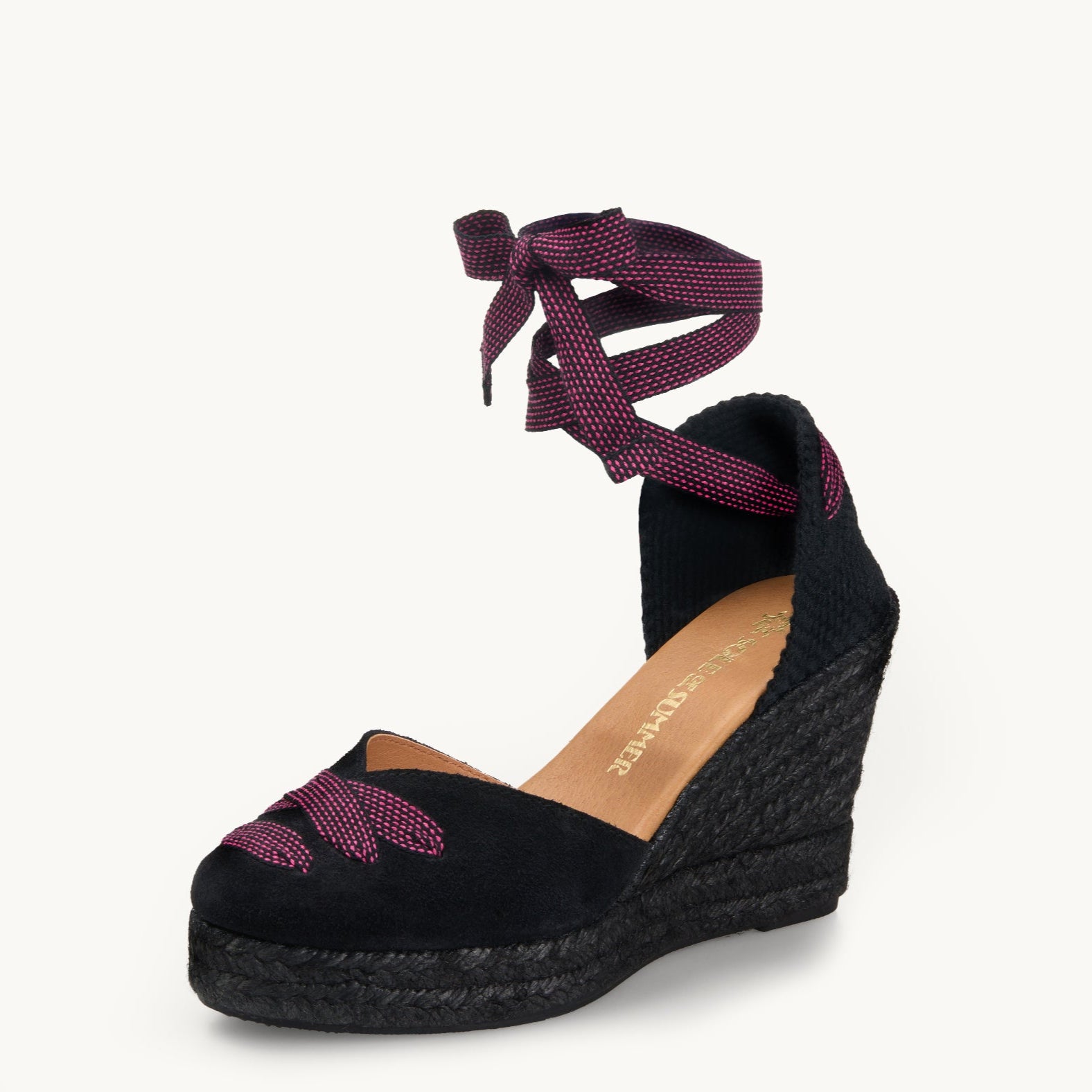 One black suede wedge espadrille displayed with black and pink dot ribbons tied in a bow as if around an ankle.  The sole is made of black Jute. The ribbon detailing is also criss-crossed over the almond shaped toe.