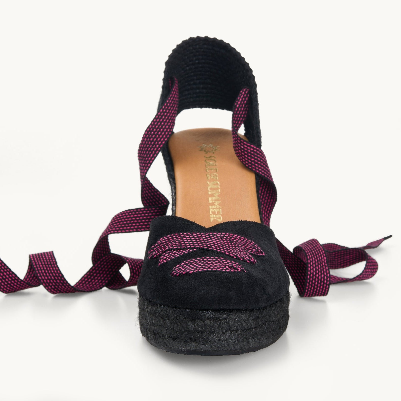 One black suede wedge espadrille displayed with black and pink dot ribbons at the heel, left loose and untied on either side. The base is made of black Jute. The ribbons are in a criss-cross detail over the almond shaped toe of the footwear. The insole is shown in tan leather with the Sole of Summer logo printed in gold.