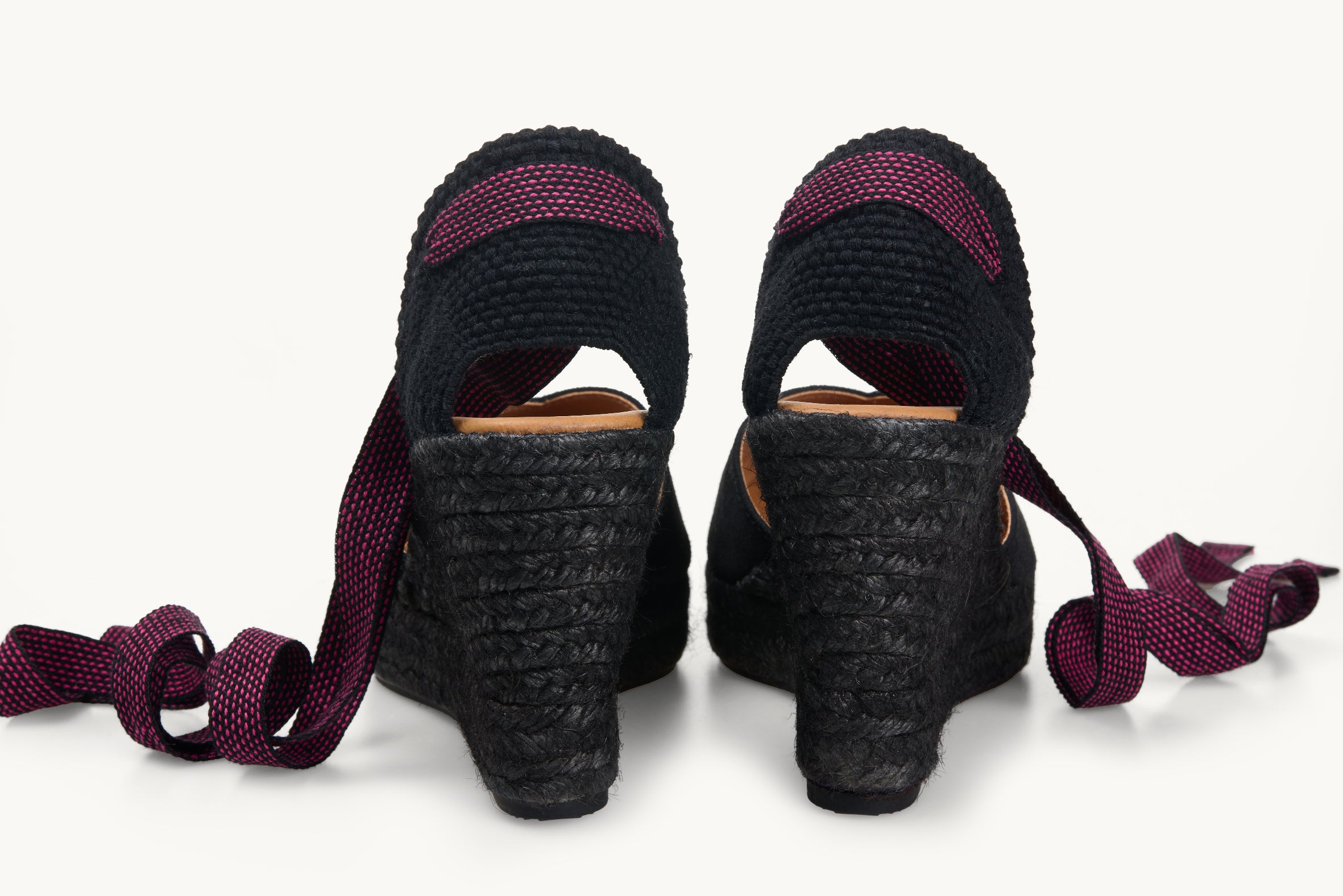 A pair of black wedge espadrilles looking at the heel details and the black jute soles are visible with the black and pink dot ribbons untied loose next to the shoes.