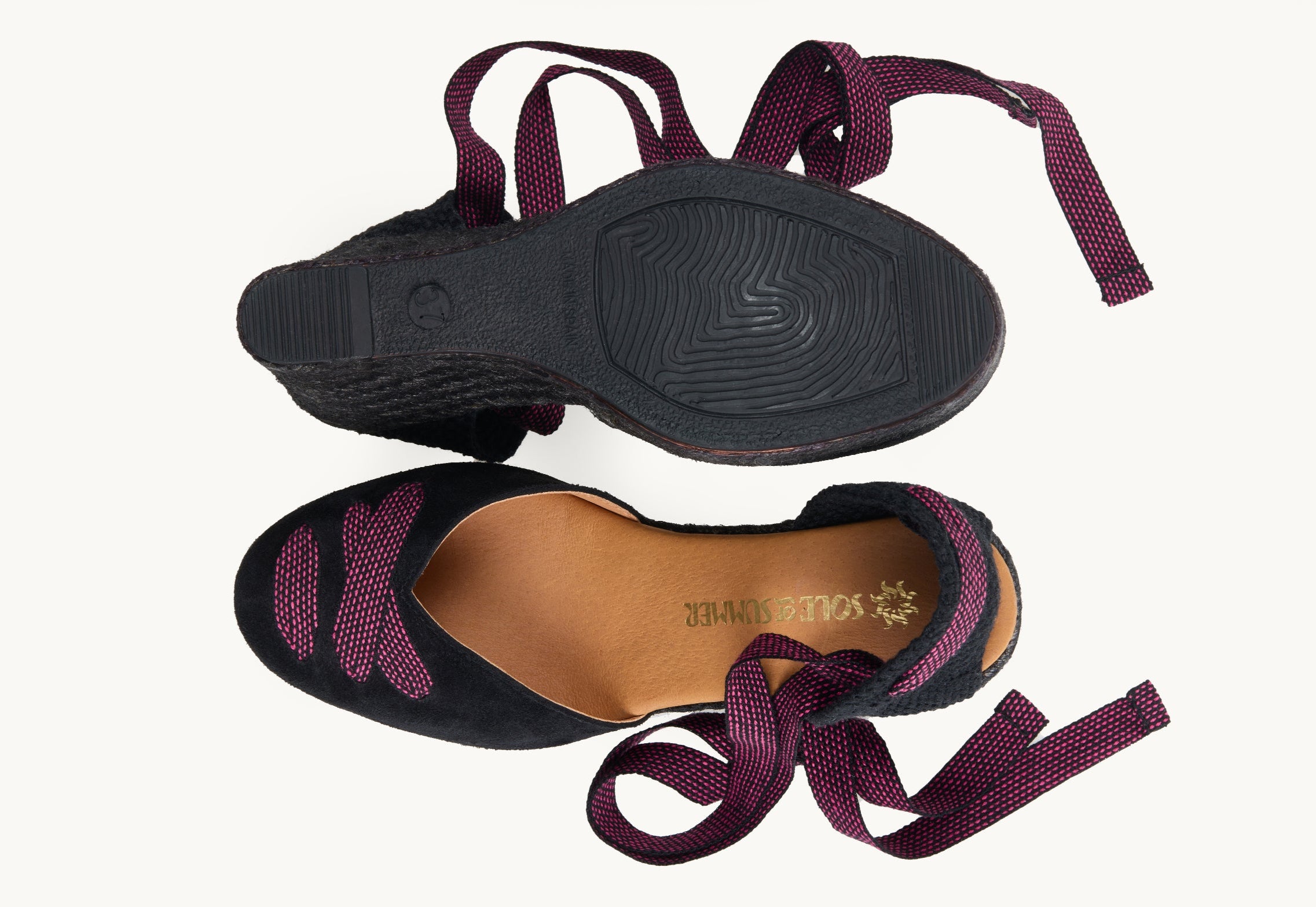 A pair of women's black suede wedge espadrilles with black and pink dot ribbons untied loose beside them. One of the footwear is turned over to show the underneath. It has a full rubber covering and the other shoe shows the design on the front of the footwear, the ribbons criss-crossed over the almond shaped toe and the inner, tan leather lined insole and the Sole of Summer logo.