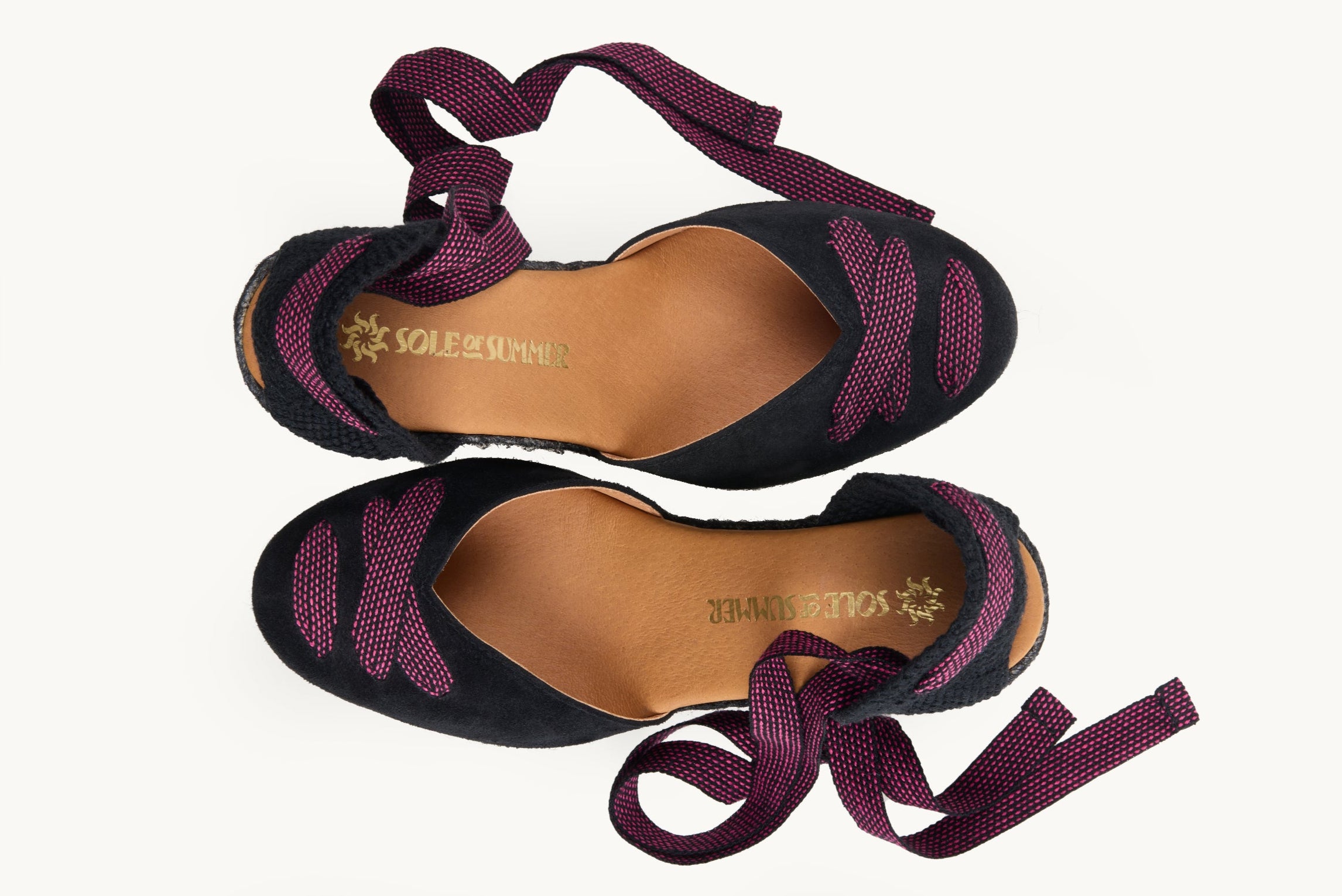 Black suede wedge espadrilles displayed as a pair with the ribbons loose and untied on either side of the shoe. View from above looking down on them. Ribbons are black with small pink dots. Both shoes have ribbon details crossed over on the almond shaped toe. View shows the inside of the footwear, tan leather lining with the Sole of Summer logo in gold.