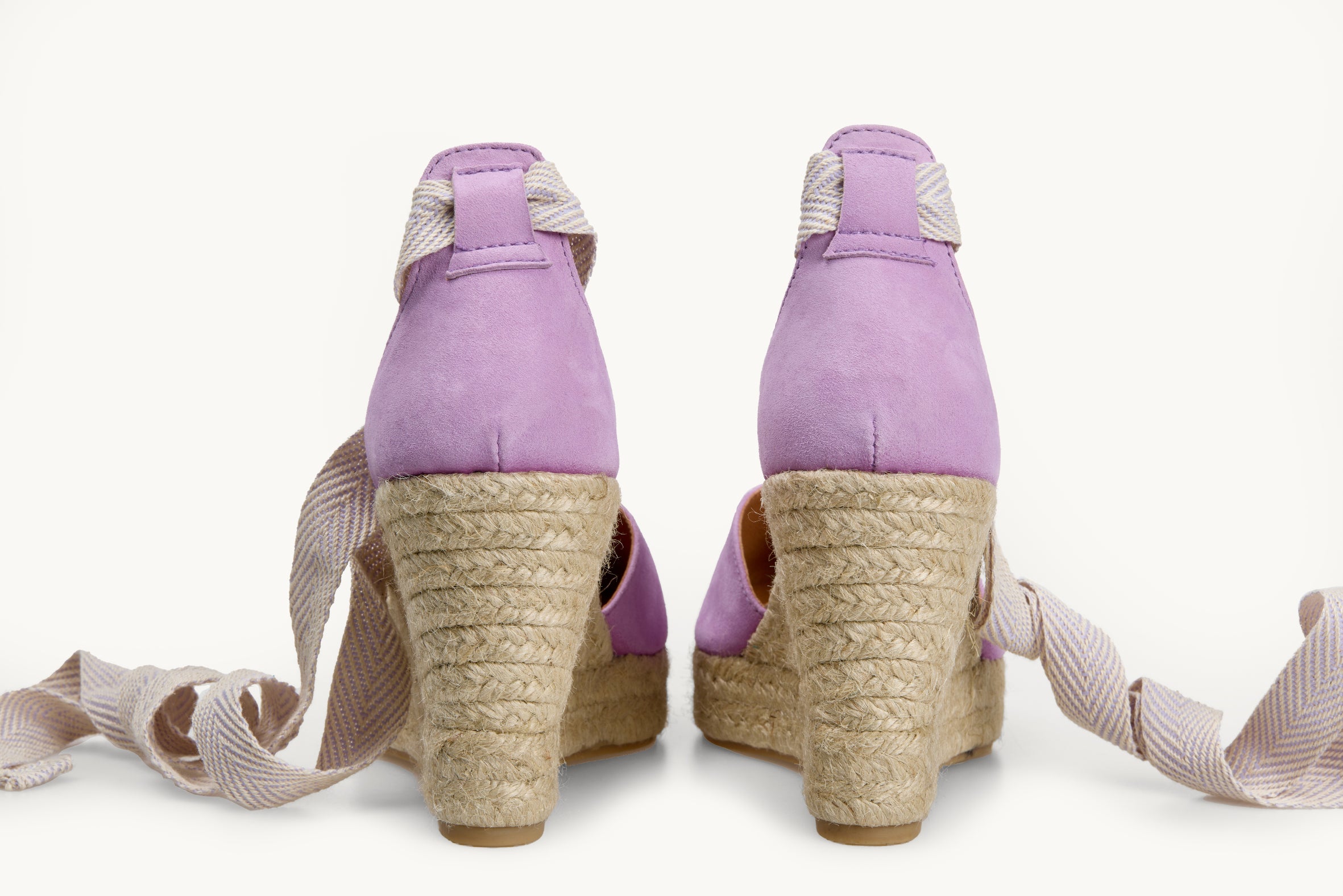 A pair of wedge espadrilles displayed with the ribbons loose untied on either side of the heels. View of the shoes from the back only of the lilac suede heels. Ribbons are white with lilac herringbone pattern. The base of the footwear is made of Jute.