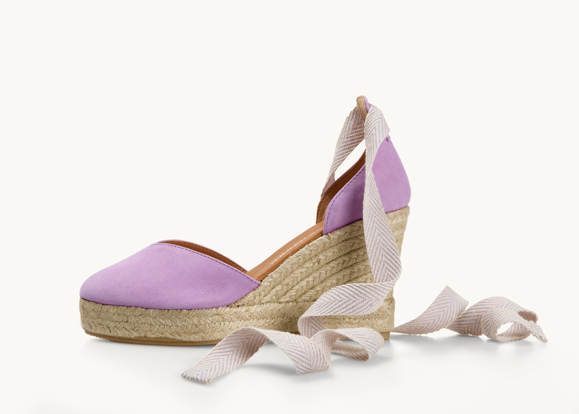 One lilac suede wedge espadrille displayed with the ribbons loose on either side of the heel of the shoe. View of the shoe from the side. Ribbons are white and lilac herringbone pattern. The base of the espadrille is made of Jute. The toe section is almond shaped. The heel is also in lilac suede.