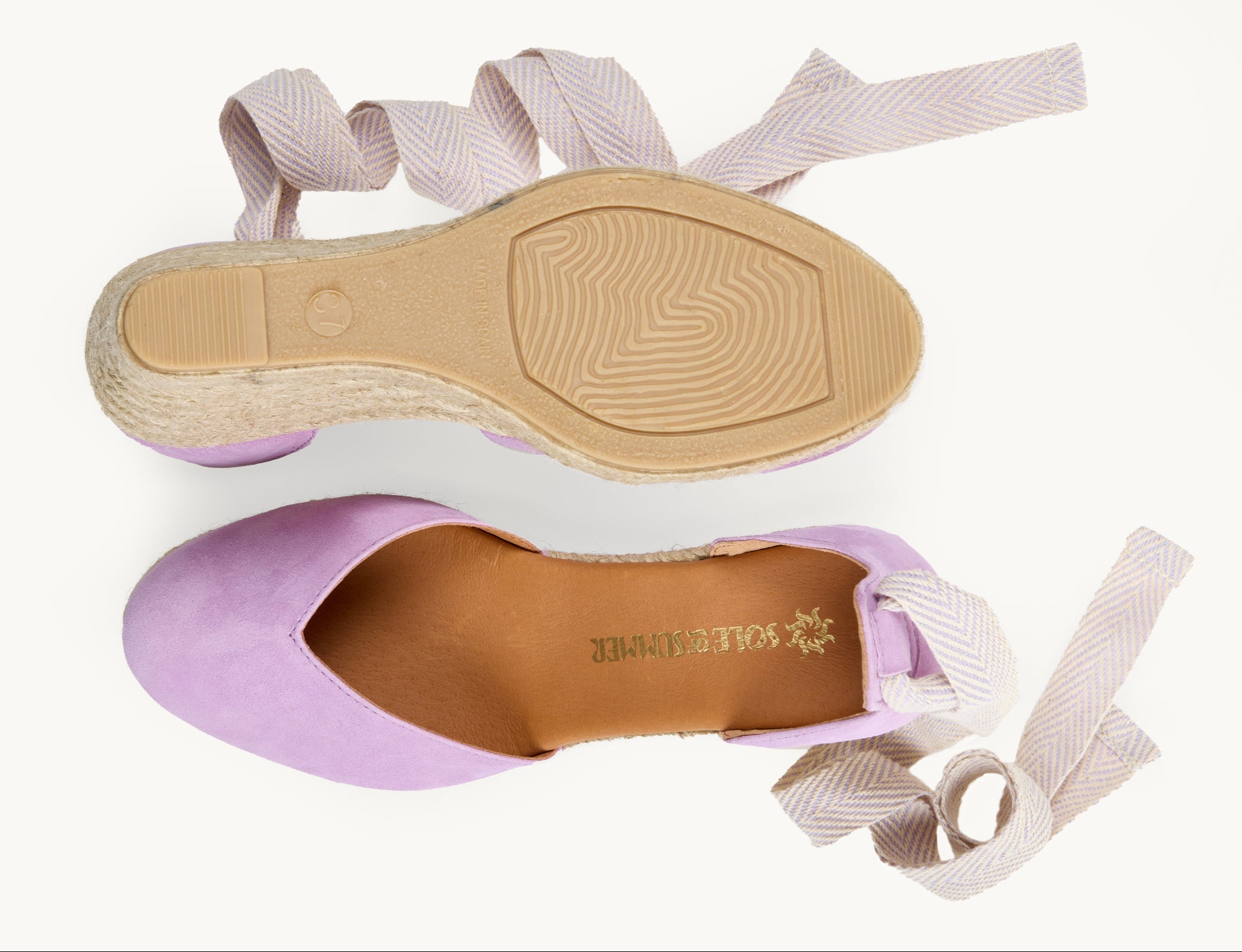 A pair of women's lilac suede wedge espadrilles with white and lilac herringbone pattern ribbons untied loose beside them. One of the footwear is turned over to show the underneath. It has a full rubber covering and the other shoe shows the inner, tan leather lined insole and the Sole of Summer logo printed in gold.