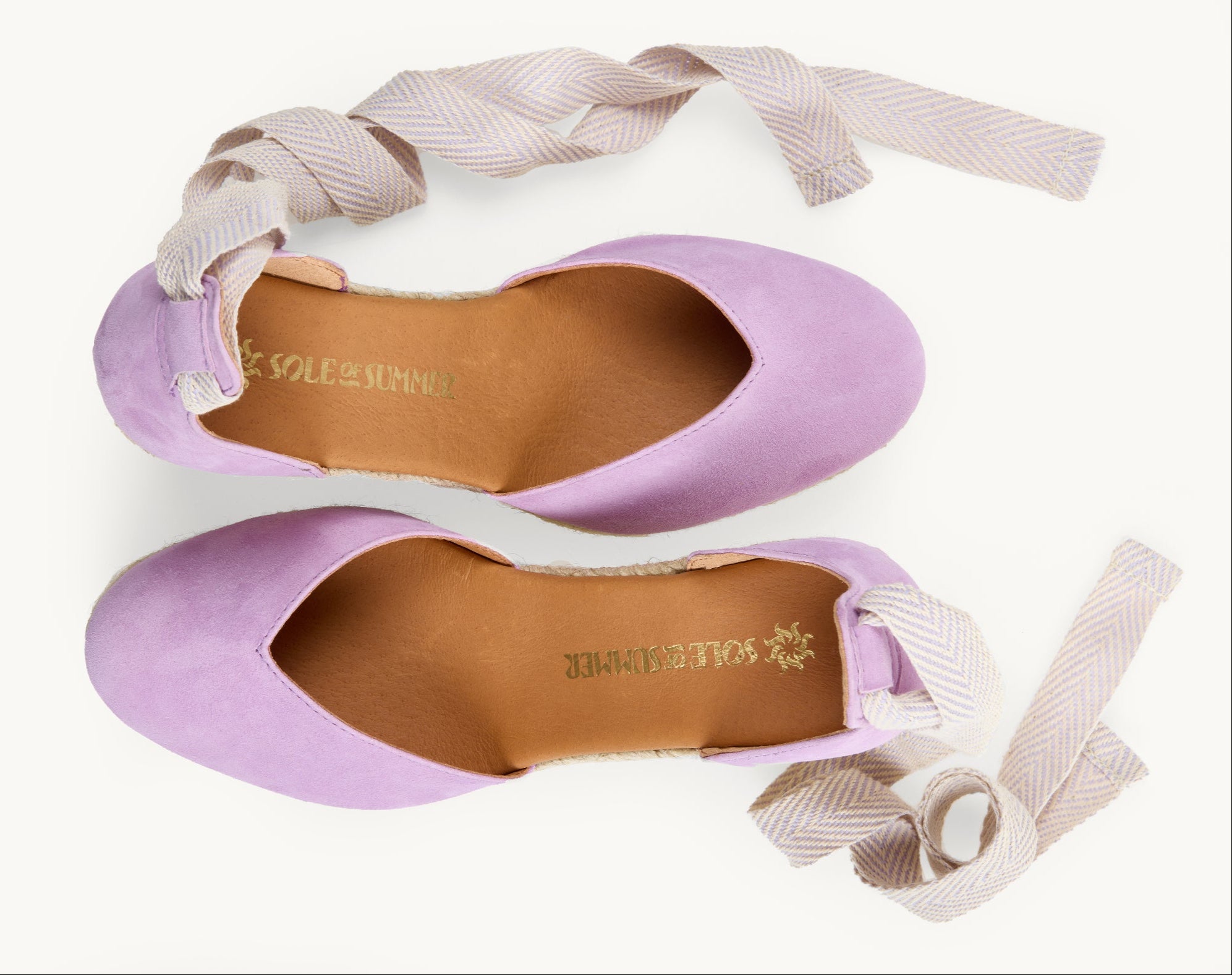 Lilac suede wedge espadrilles displayed as a pair with the ribbons loose and untied on either side of the shoe. View from above looking down on them. Ribbons are white and lilac herringbone pattern. View shows the inside of the footwear, tan leather lining with the Sole of Summer logo printed in gold.