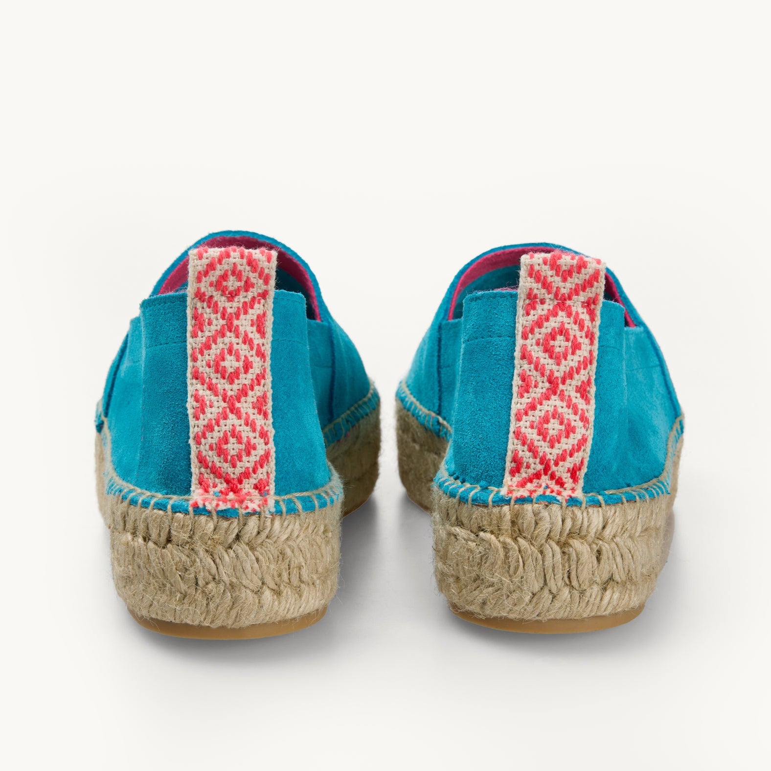 A pair of turquoise suede women's espadrille loafers. Only the back details of the heels are shown. A stripe of neon pink and white patterned ribbon about a centimetre wide runs straight up the back heel. Stitching around the edge of the upper to join it to the traditional jute sole.