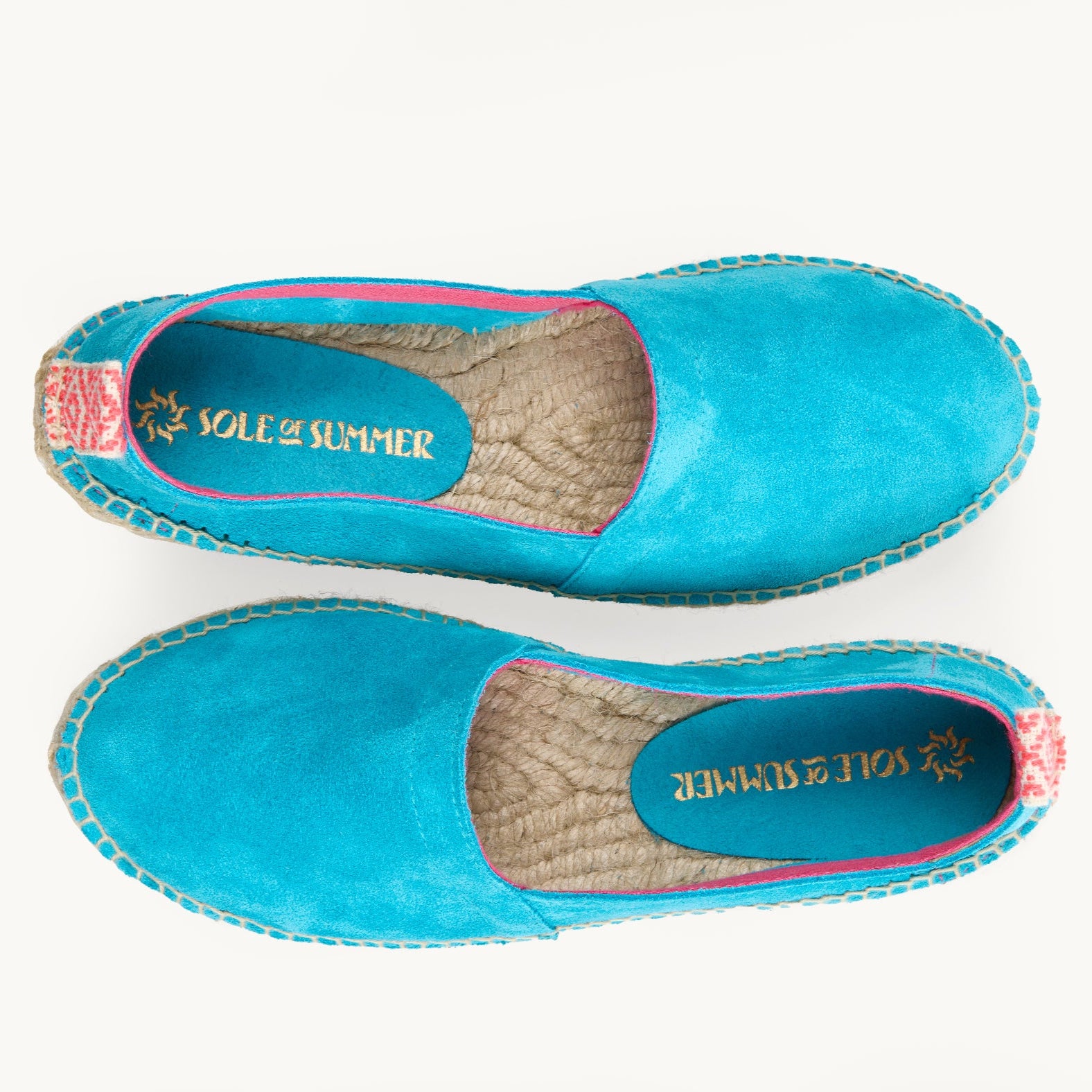 A pair of loafer espadrilles. Flat shoes. Arial view. Turquoise suede upper with pink trim around the opening with stitching around the edge joining the upper to the traditional jute sole. Pink and white diamond pattern ribbon up the heel of the loafer.