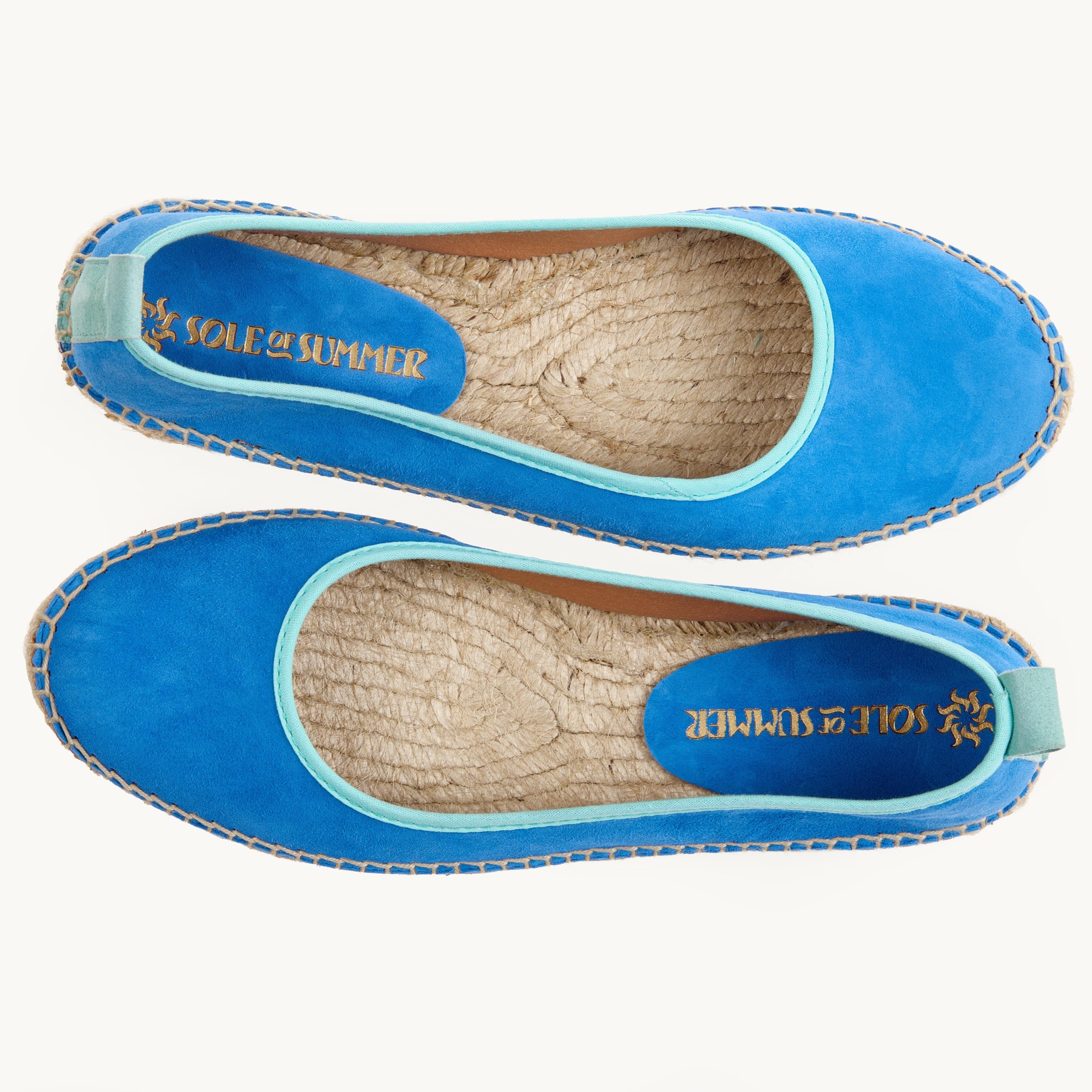 A pair of flat ballet espadrilles. View from above looking down on the footwear, unlined jute inside is shown. Azure blue suede upper with duck egg/light turquoise cotton trim around the opening of the shoe with stitching around the edge joining the upper to the a traditional jute sole.