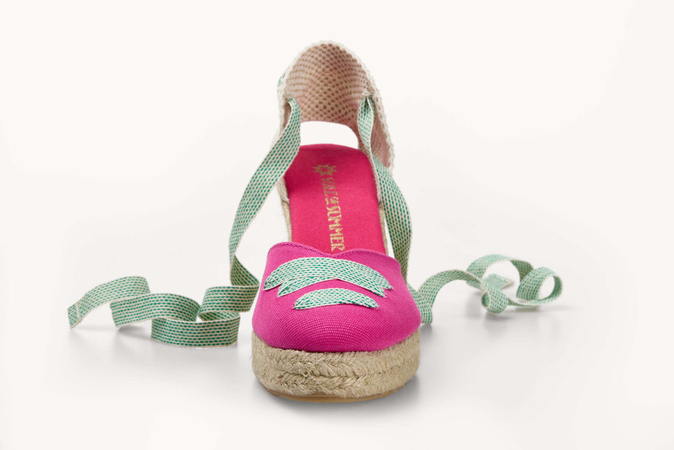 One bright pink cotton wedge espadrille displayed with white and green dot ribbons at the heel, left loose and untied on either side. The base is made of Jute. The ribbons are in a criss-cross detail over the almond shaped toe of the footwear.