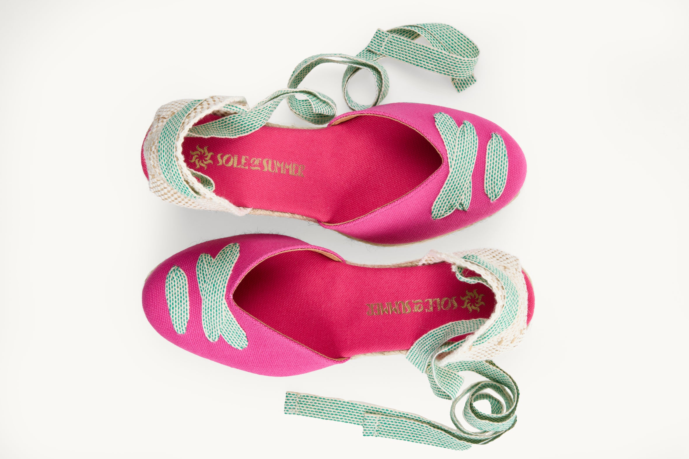 Bright pink cotton wedge espadrilles displayed as a pair with the ribbons loose and untied on either side of the shoe. View from above looking down on them. Ribbons are white with small green dots. Both shoes have ribbon details crossed over on the almond shaped toe. View shows the inside of the footwear, bright pink lining with the Sole of Summer logo in gold.