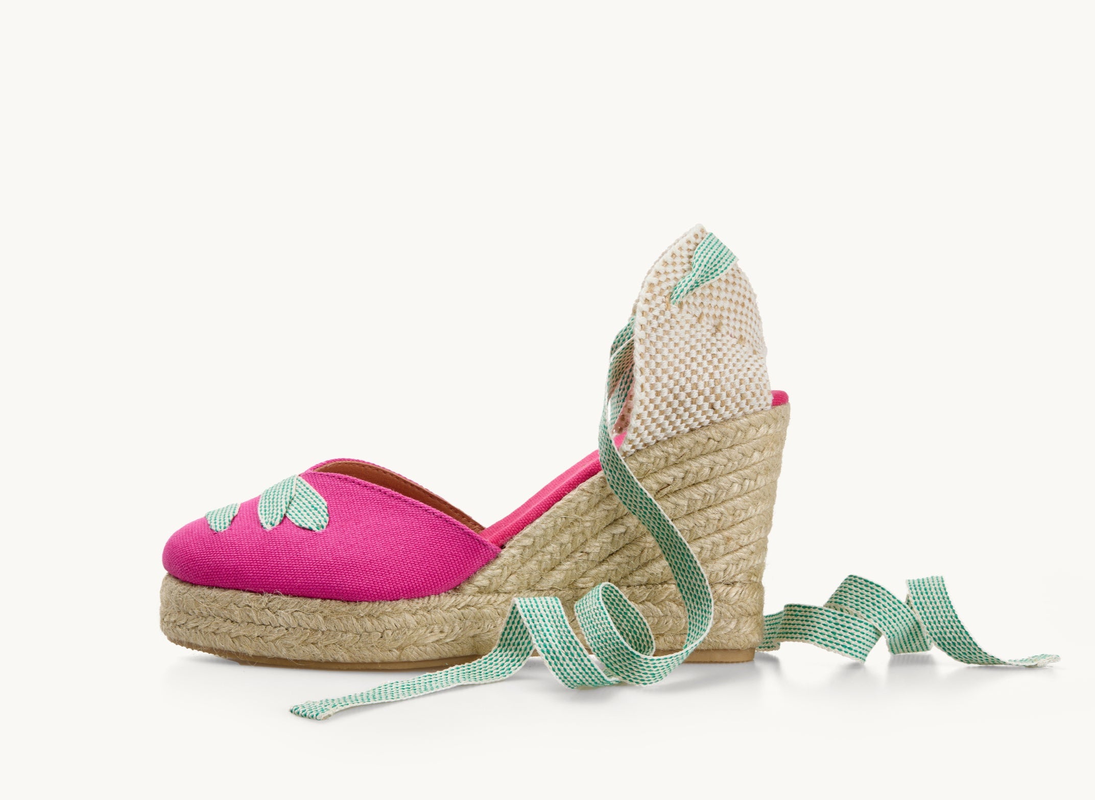 One bright pink cotton wedge espadrille shoe, side view. It has a jute sole. Across the almond shaped toe is a criss-cross of white ribbon with green dot pattern. The ankle ties are in the same ribbon material and are untied and loose to the side.