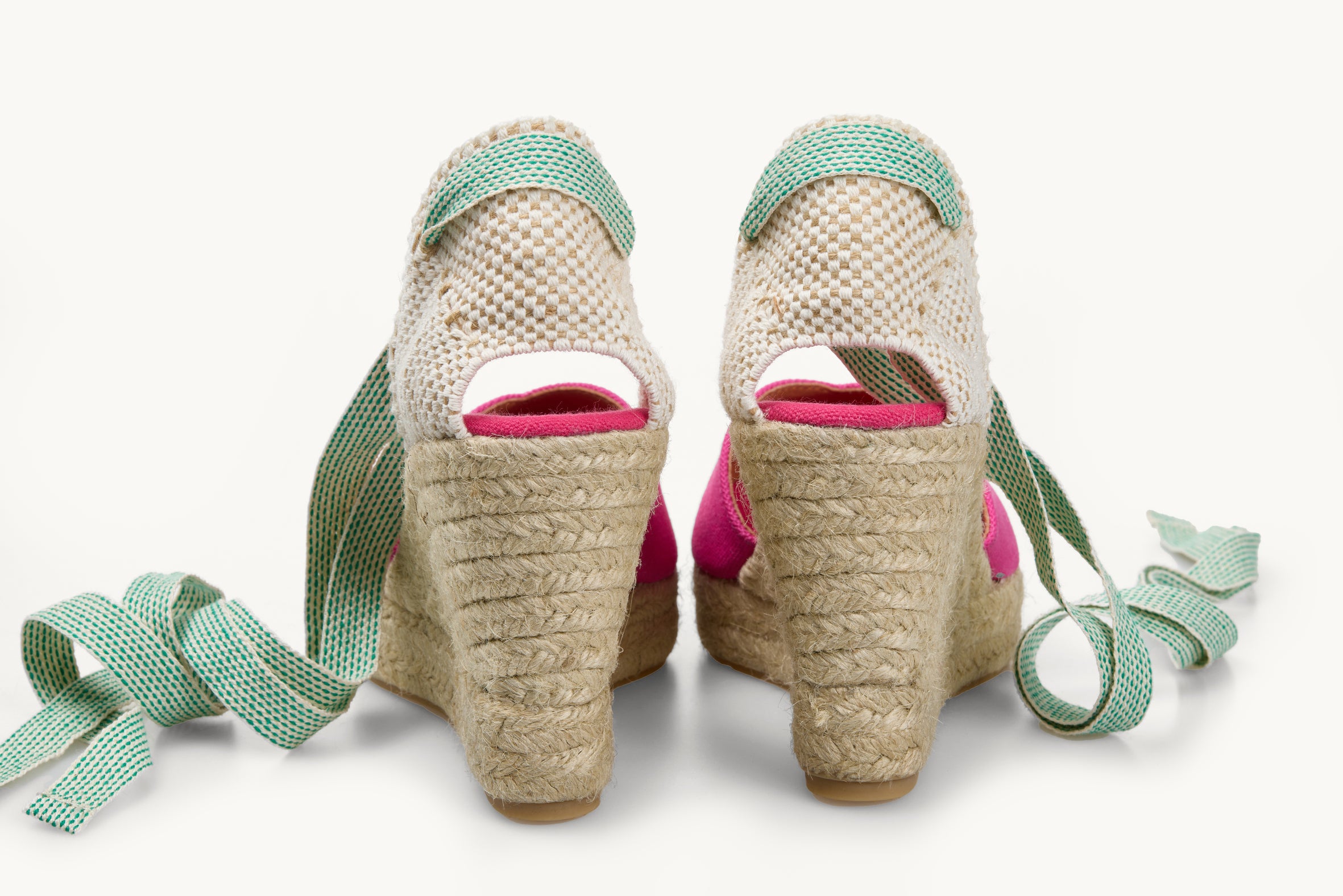 A pair of pink wedge espadrilles looking at the heel details and the jute soles are visible with the white and green dot ribbons untied loose next to the shoes.