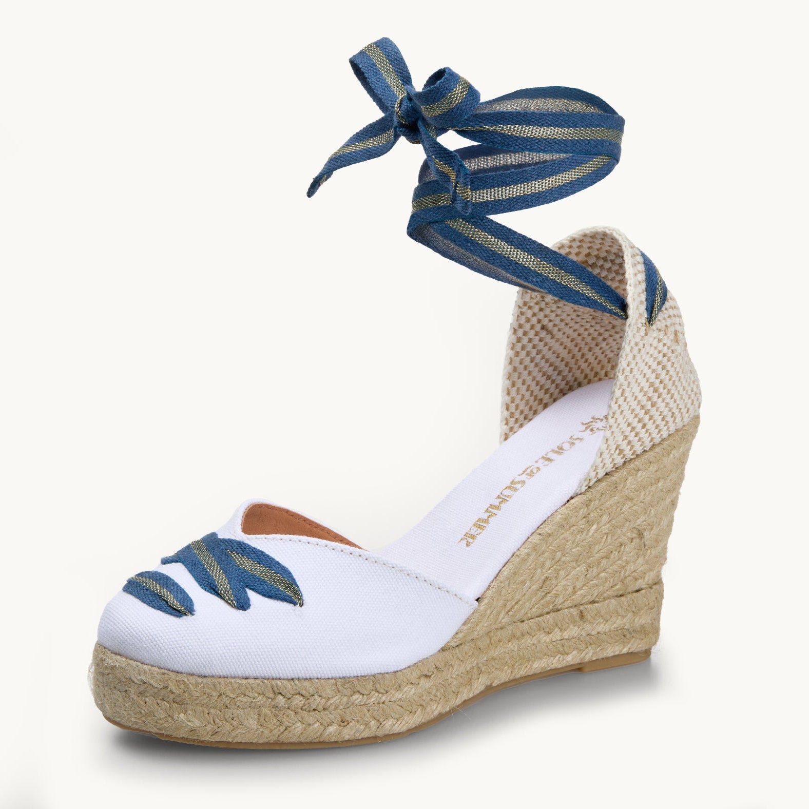 One white cotton wedge espadrille displayed with blue and gold stripe ribbons tied in a bow as if around an ankle.  The sole is made of Jute. The ribbon detailing is also criss-crossed over the almond shaped toe.