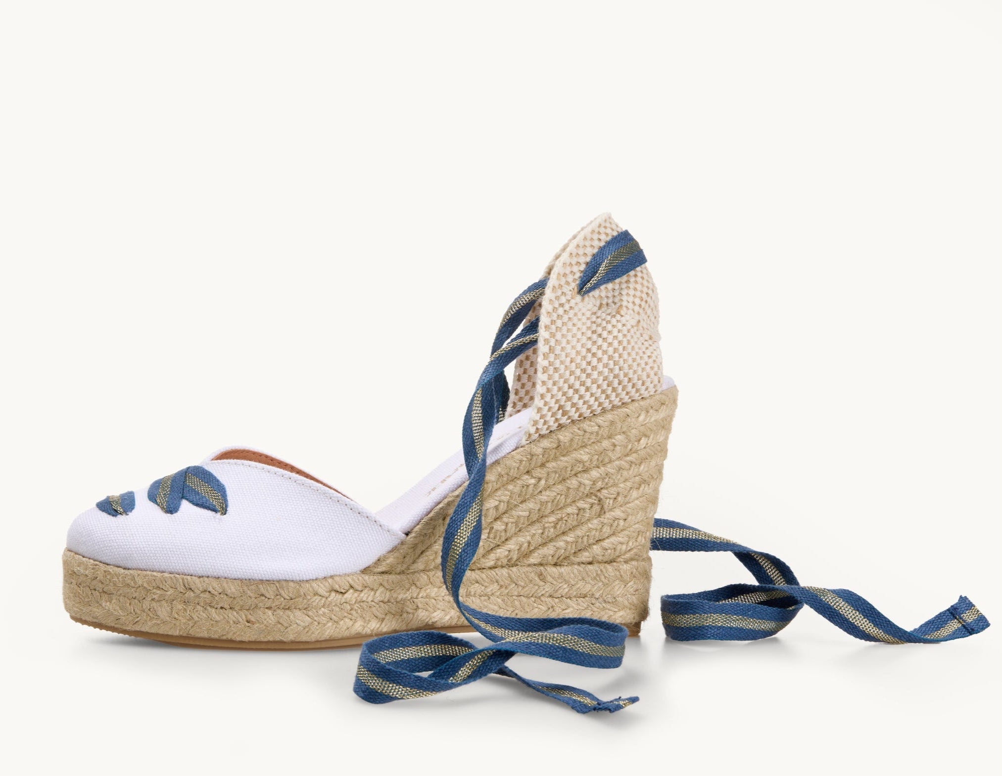 One white cotton wedge espadrille displayed with the ribbons loose on either side of the heel of the shoe. View of the shoe from the side. Ribbons are blue and gold striped. The base of the espadrille is made of Jute. Blue and gold ribbon detail on the almond shaped toe of the footwear.