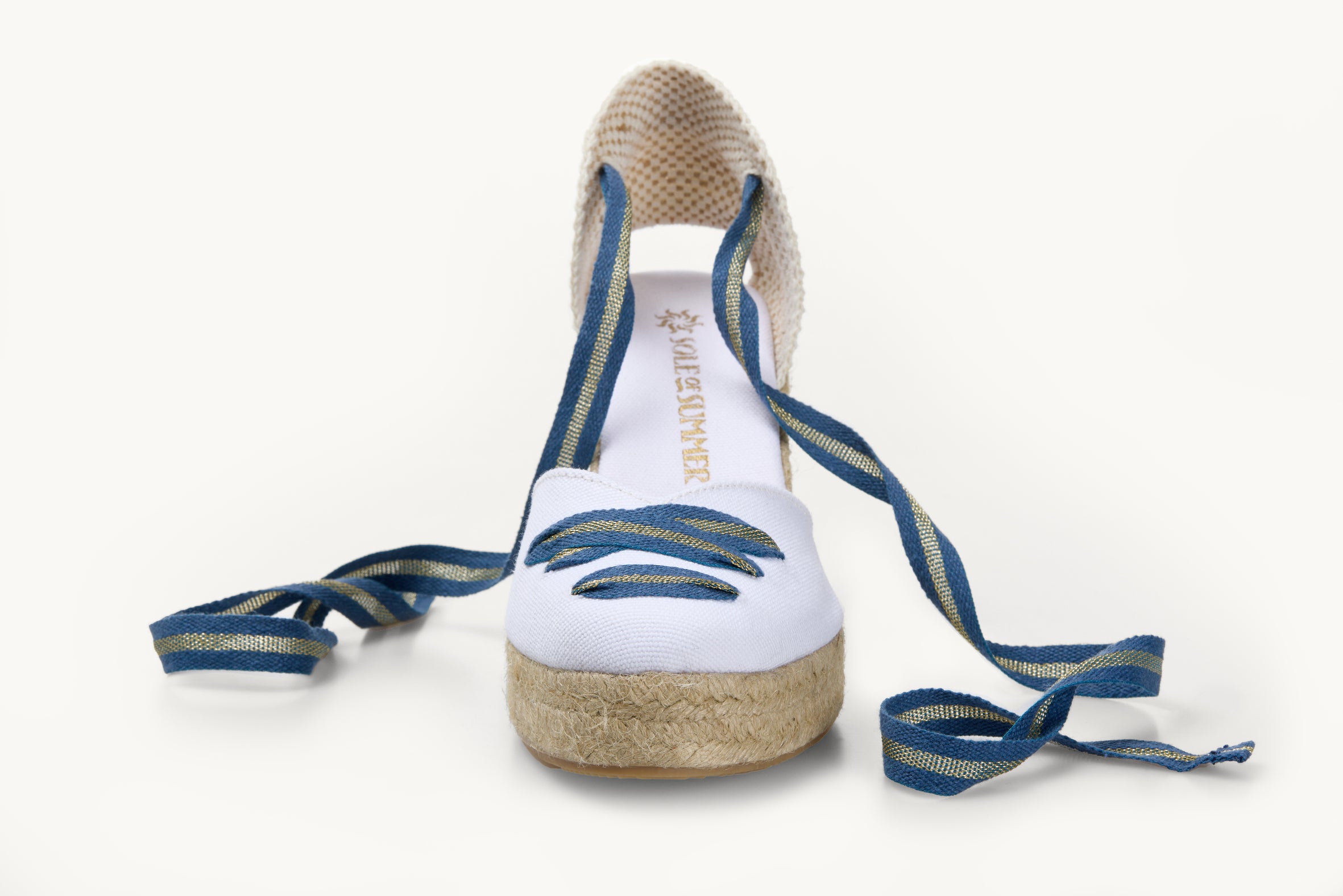 One white cotton wedge espadrille displayed with blue and gold stripe ribbons at the heel, left loose and untied on either side. The base is made of Jute. The ribbons are in a criss-cross detail over the almond shaped toe of the footwear.