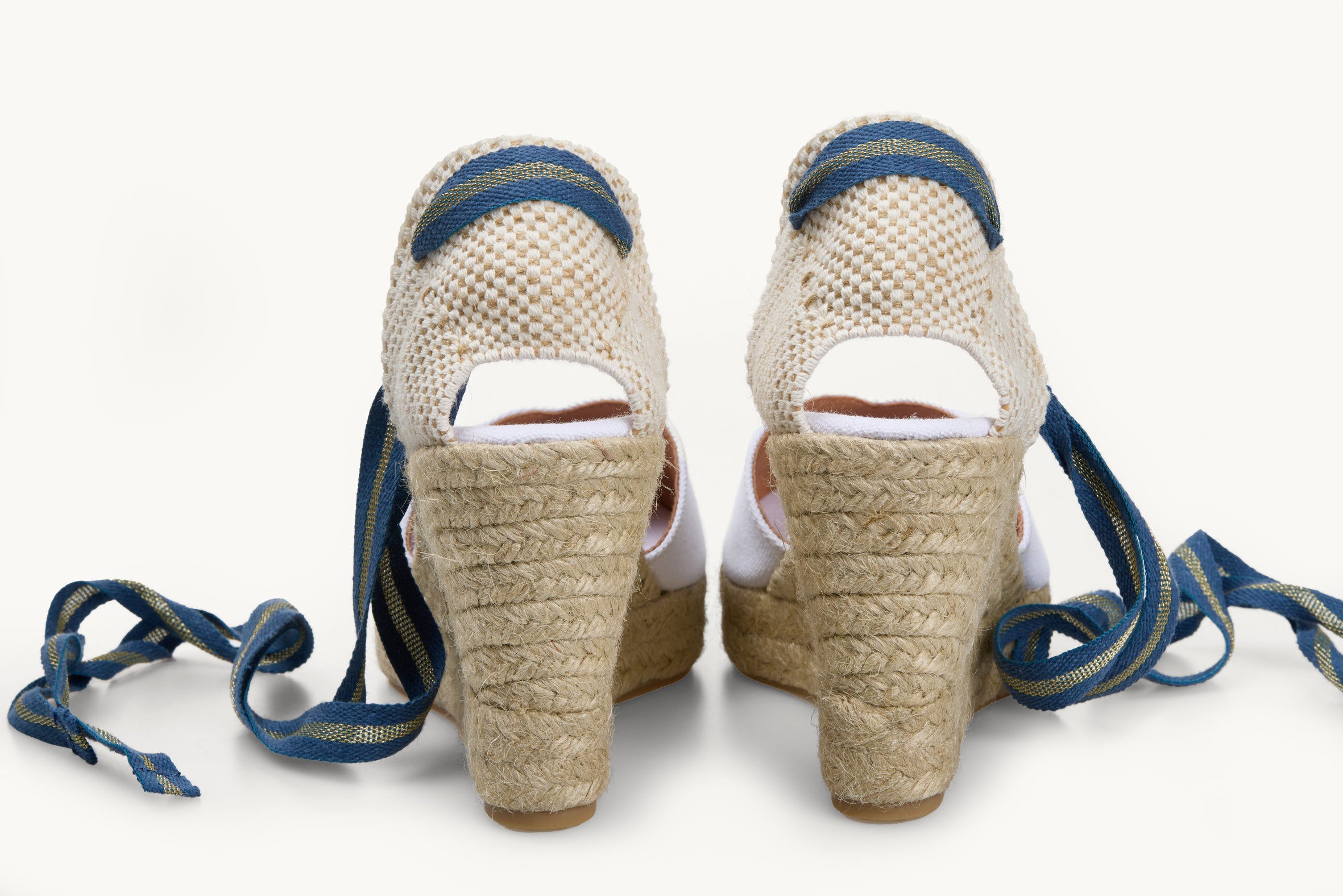 A pair of wedge espadrille displayed with the ribbons loose untied on either side of the heels. View of the shoes from the back only of the heels. Ribbons are blue and gold striped. The base of the footwear is made of Jute.