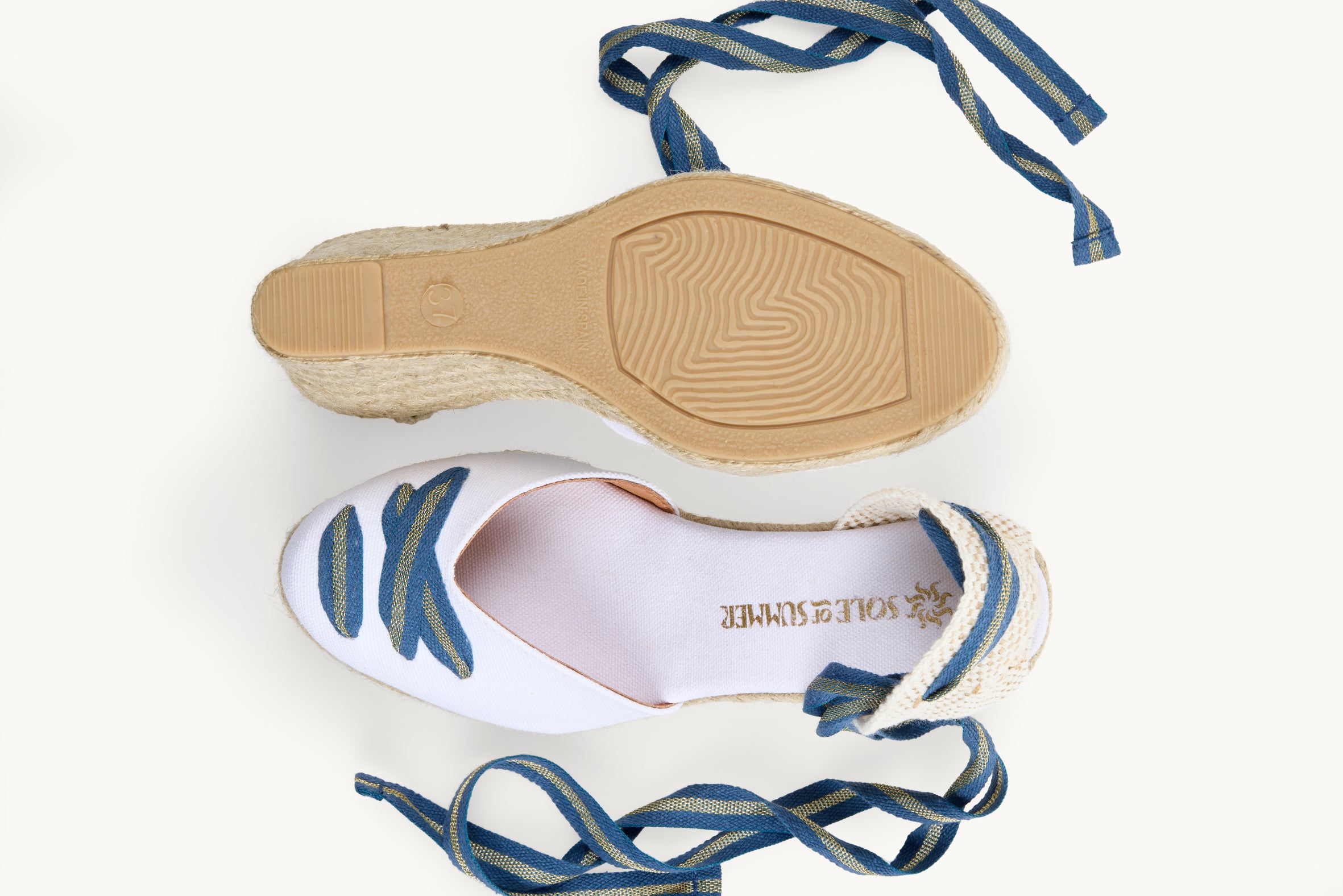 A pair of women's white cotton wedge espadrilles with blue and gold striped ribbons untied loose beside them. One of the footwear is turned over to show the underneath. It has a full rubber covering and the other shoe shows the inner, white lined insole and the Sole of Summer logo.
