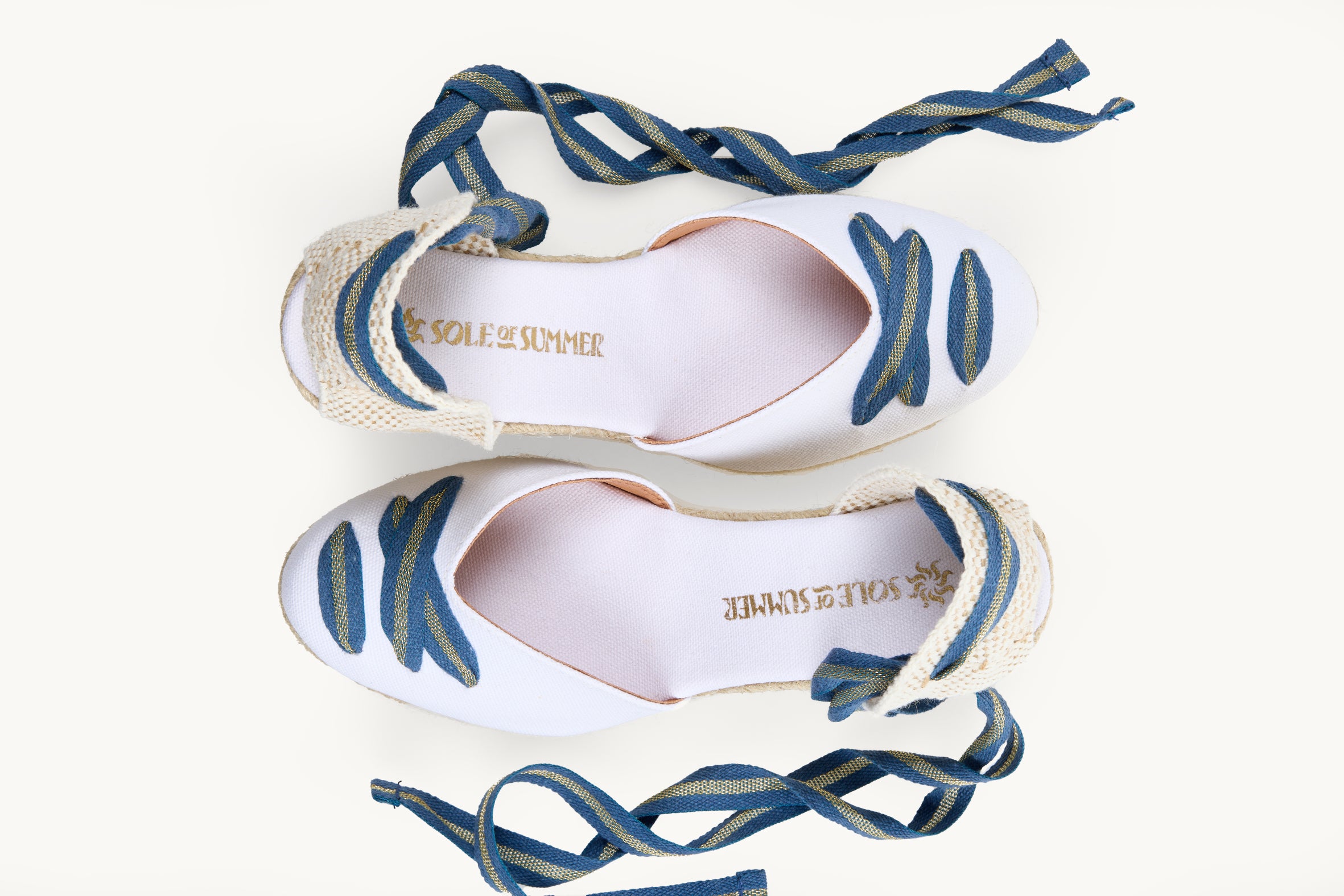 White cotton wedge espadrilles displayed as a pair with the ribbons loose and untied on either side of the shoe. View from above looking down on them. Ribbons are blue and gold striped. Both shoes have ribbon details crossed over on the almond shaped toe. View shows the inside of the footwear, white lining with the Sole of Summer logo in gold.