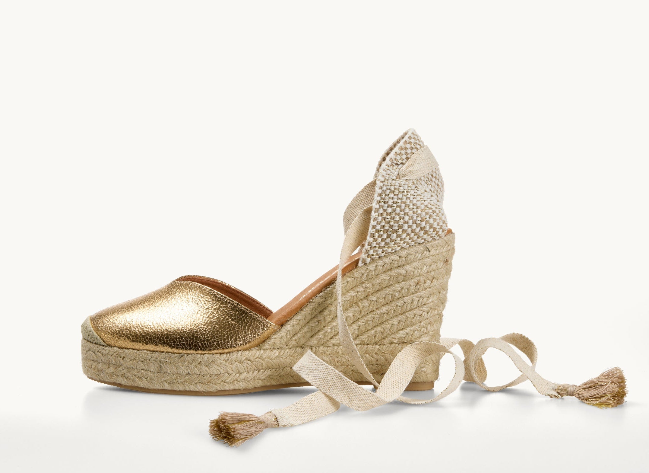 One crackled gold leather wedge espadrille displayed with the ribbons loose on either side of the heel of the shoe. View of the shoe from the side. Ribbons are cream with gold tassels on the ends. The base of the espadrille is made of Jute. The toe is almond shaped.