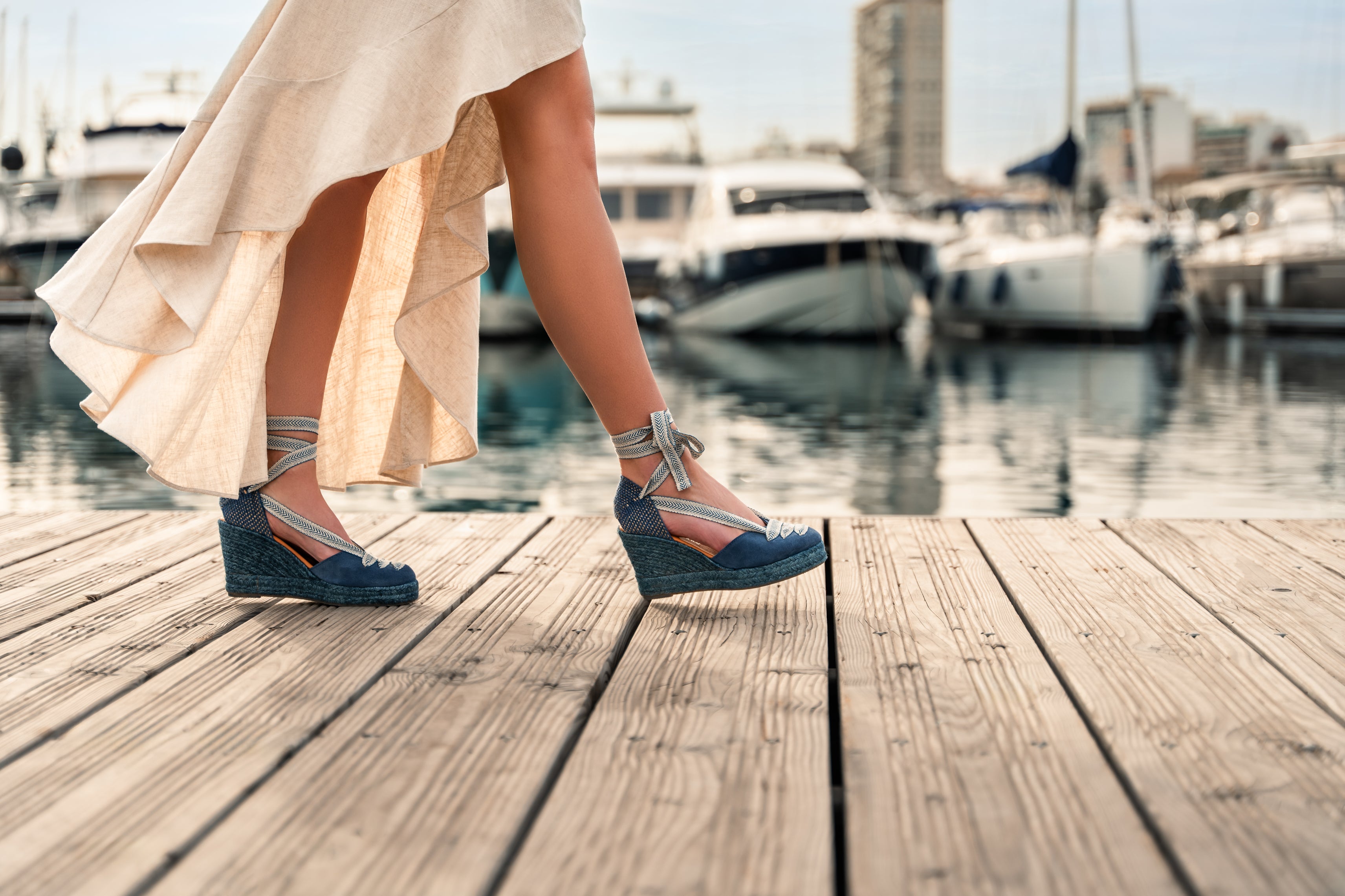 women walking along a wooden deck with yachts in the background. Wearing a flowing ream dress and blue wedge espadrilles with blue soles and ribbons tied up her ankles.