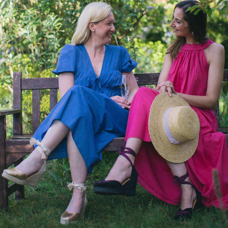 Two women sitting on a bench in a garden, one in a blue dress and the other in a pink dress with a hat.