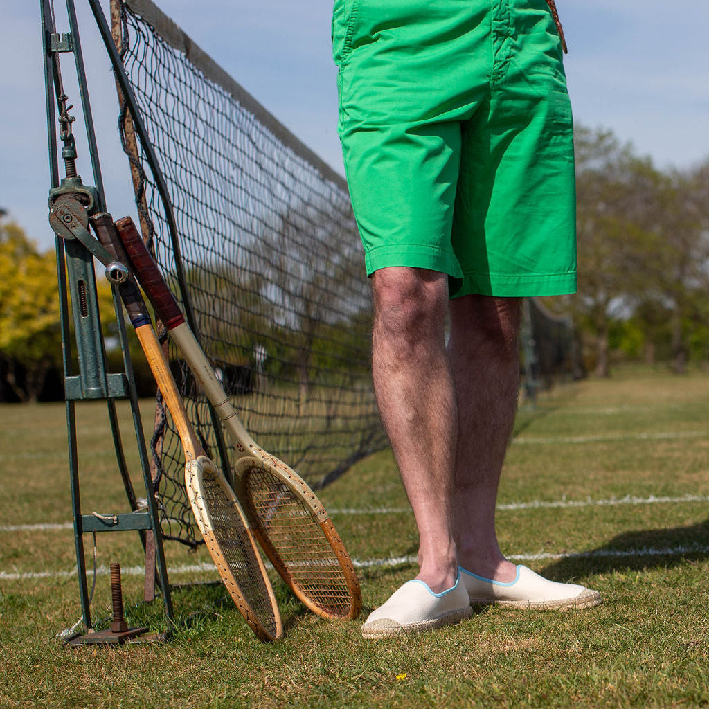 Person wearing green shorts standing on a grassy tennis court with vintage rackets leaning against the net wearing white shoes.