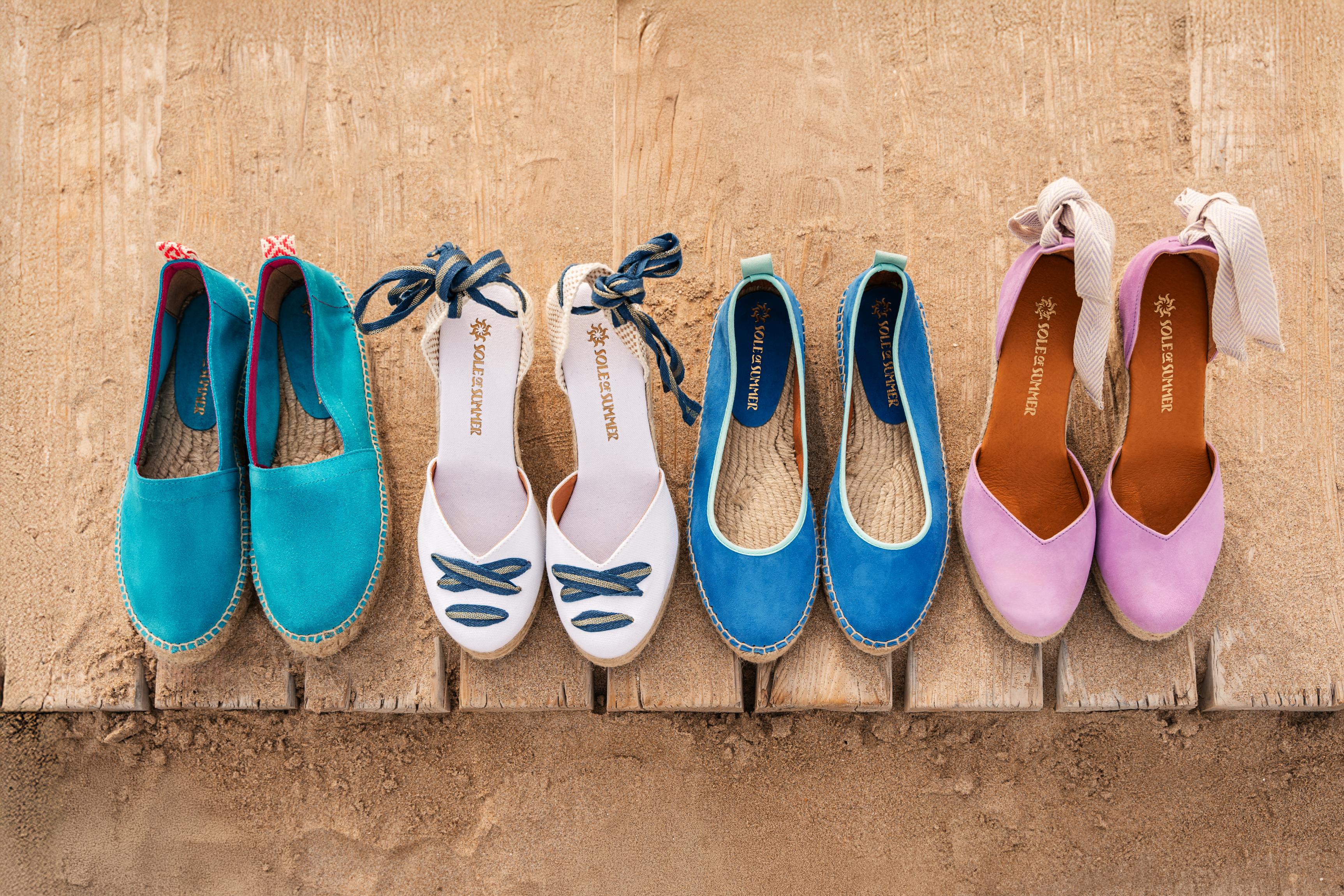 4 pairs of women's espadrilles lined up on a sandy wooden path at the beach. Turquoise loafers, white wedge, blue ballet flats and lilac suede wedge espadrilles.