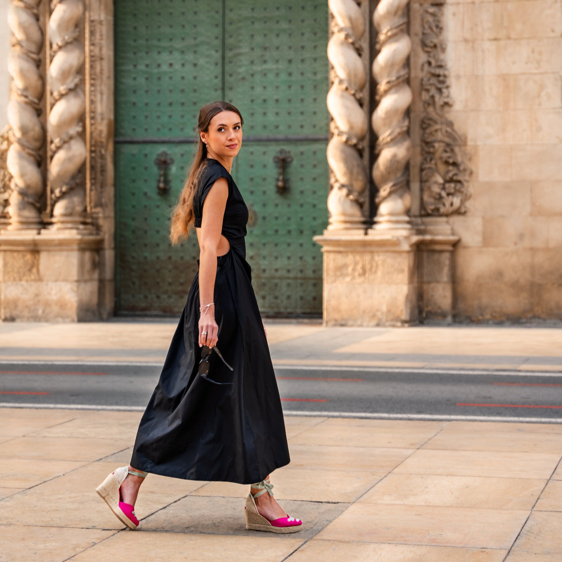 A women in a black dress holding sunglasses is walking past a grand old doorway with green doors. She is wearing pink espadrille wedge shoes with ribbons tied around her ankles in bows.