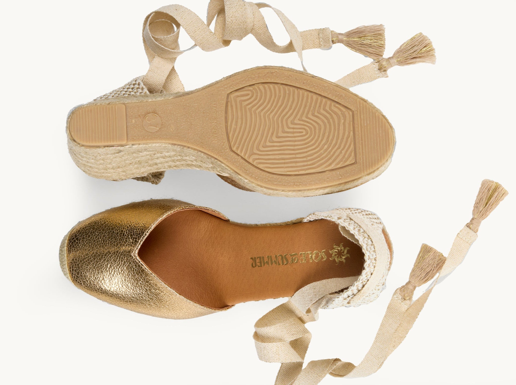 A pair of women's crackled leather in gold wedge espadrilles with cream ribbons with gold tassels on the ends untied loose beside them. One of the footwear is turned over to show the underneath. It has a full rubber covering and the other shoe shows the inner, tan leather lined insole and the Sole of Summer logo.