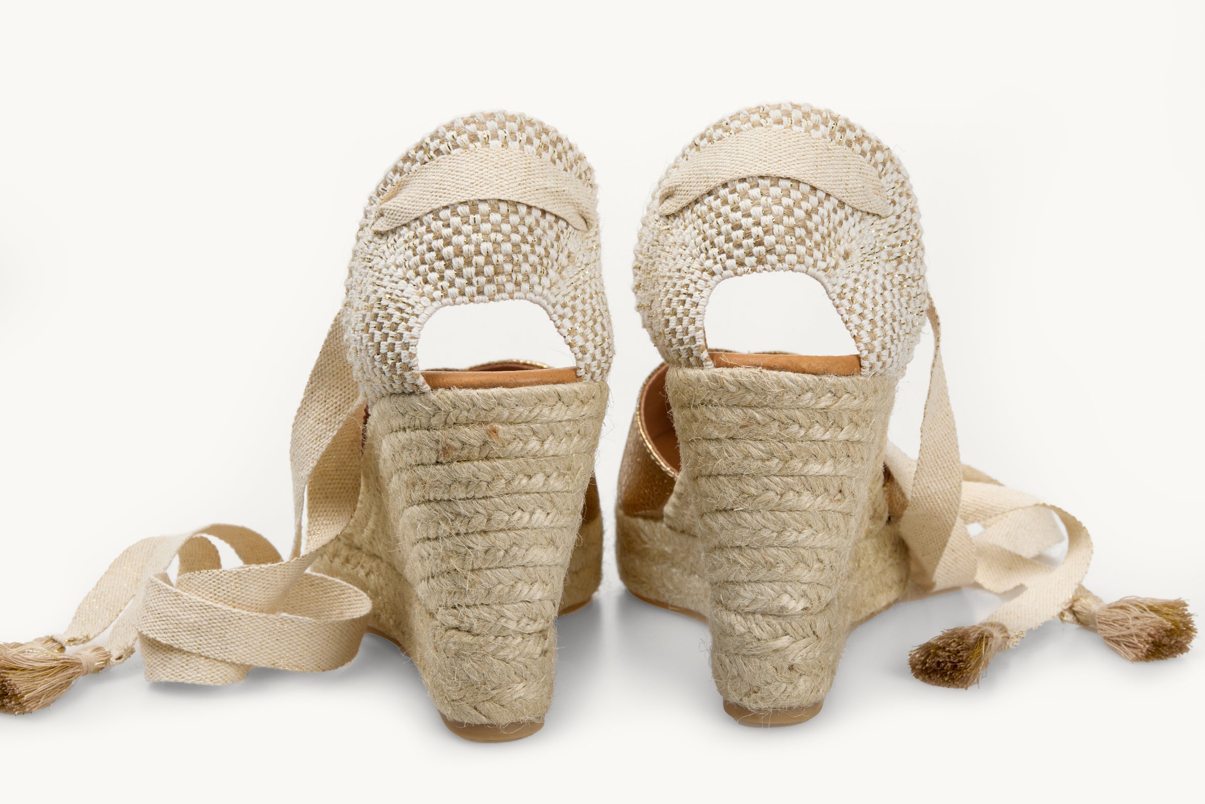 A pair of wedge espadrille displayed with the ribbons loose untied on either side of the heels. View of the shoes from the back only of the heels. Ribbons are cream with gold tassels on the ends. The base of the footwear is made of Jute.