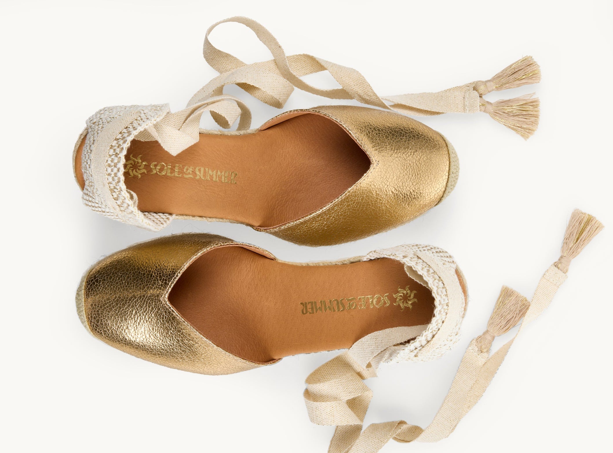 Crackled gold leather wedge espadrilles displayed as a pair with the ribbons loose and untied on either side of the shoe. View from above looking down on them. Ribbons are cream with gold tassels on the ends. The toes are almond shaped. View shows the inside of the footwear, tan leather lining with the Sole of Summer logo in gold.