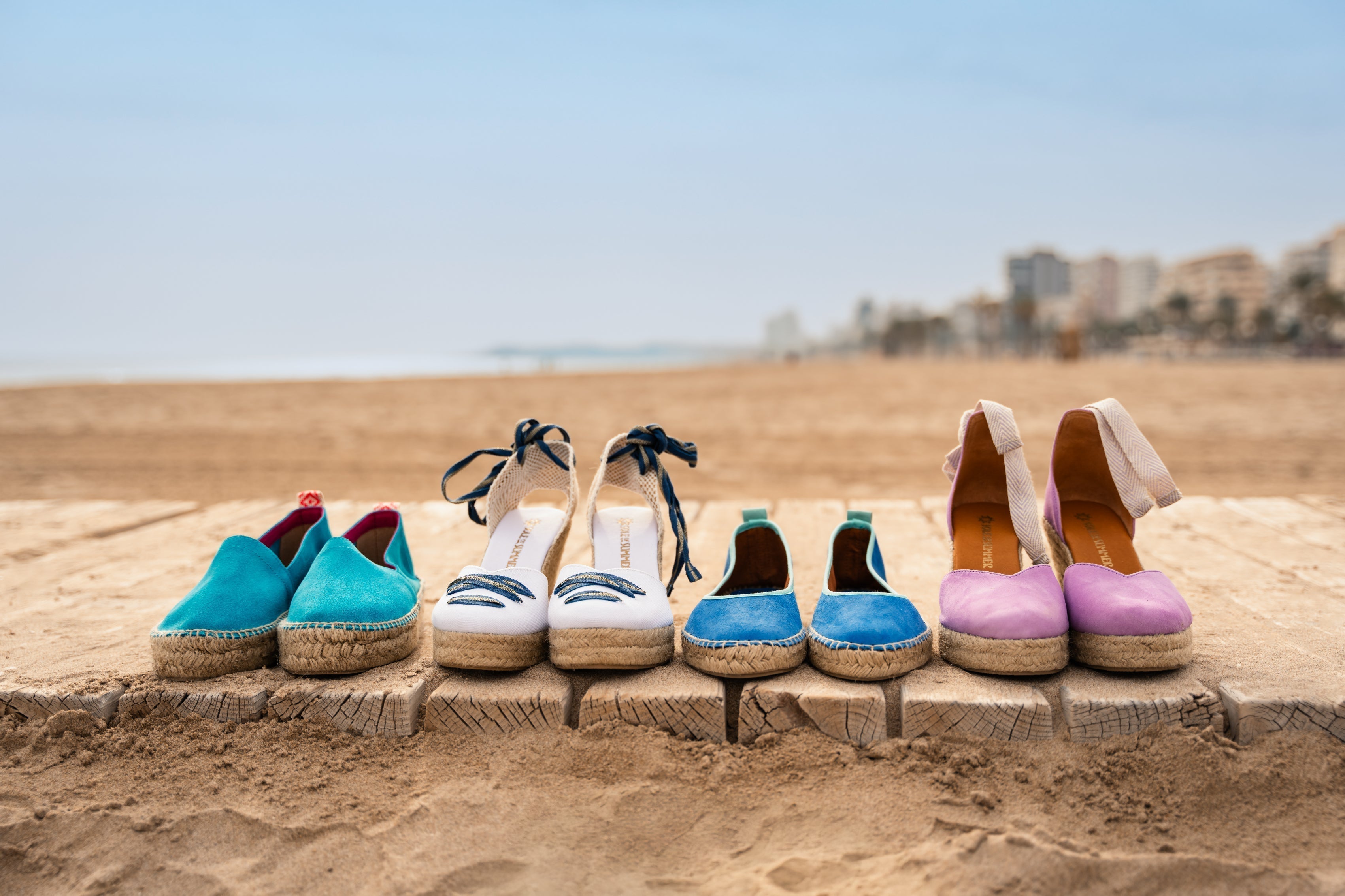 Image shows 4 pairs of ladies espadrilles shoes in turquoise, white, blue and lilac. Lined up on a boardwalk on a beach surrounded by sand with a blue sky and buildings in the horizon.