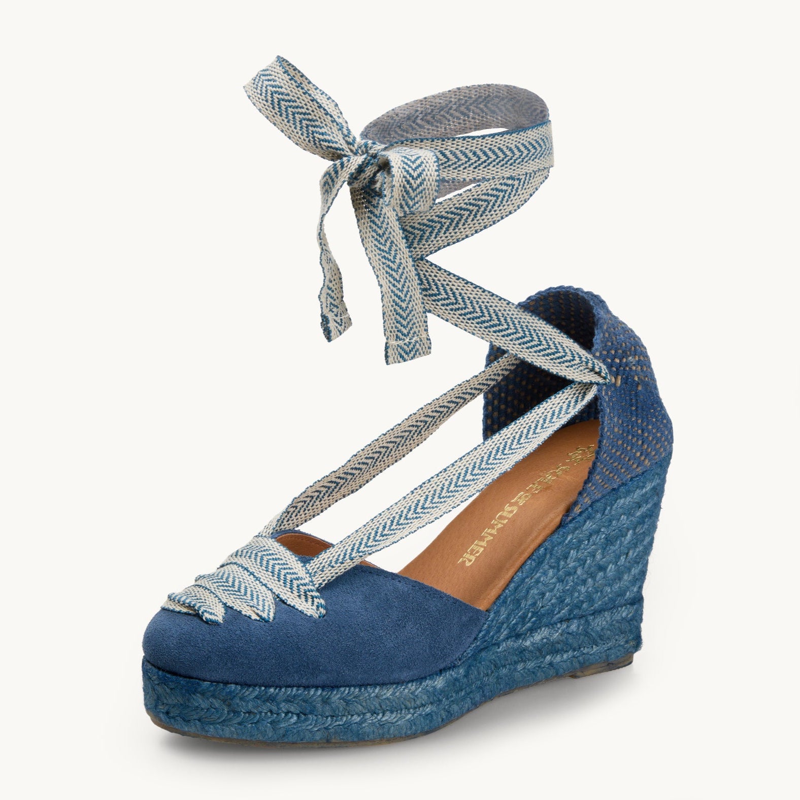 One iris denim blue suede wedge espadrille displayed with blue and white arrow head pattern ribbons tied in a bow as if around an ankle.  The sole is made of matching blue Jute. The ribbon detailing over the almond shaped toe goes across in three lines. There are two ribbons going from the toe to the heel on the left and right of the footwear.