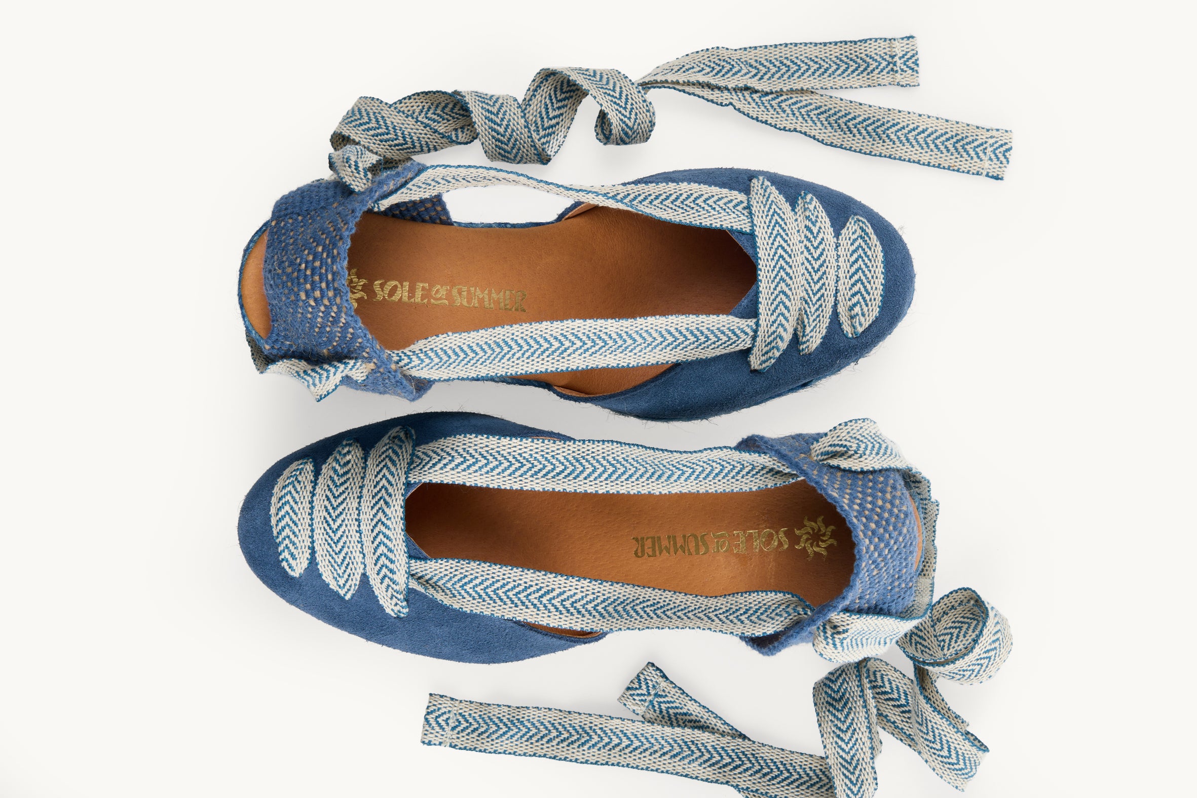 Iris denim blue suede wedge espadrilles displayed as a pair with the ribbons loose and untied on either side of the shoe. View from above looking down on them. Ribbons are blue and white arrow head pattern.  Both shoes have ribbon details in three rows over the almond shaped toe. View shows the inside of the footwear, tan leather lining with the Sole of Summer logo in gold.