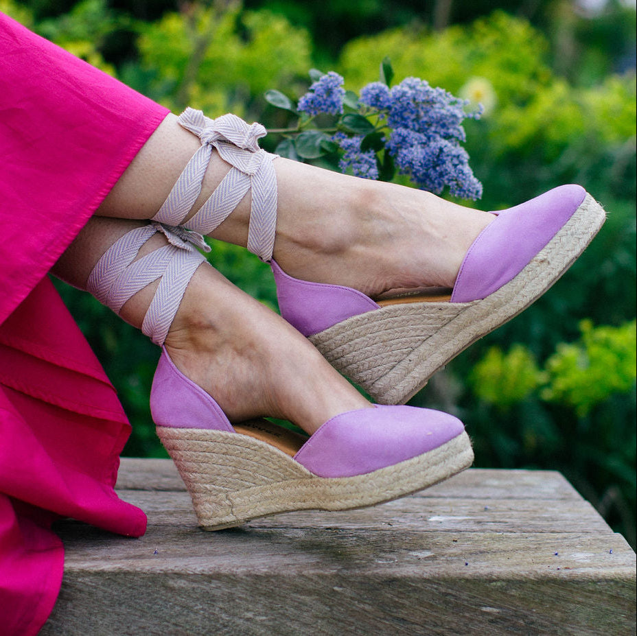 Purple wedge sandals with ribbons worn by a person in a pink skirt against a green outdoor background.