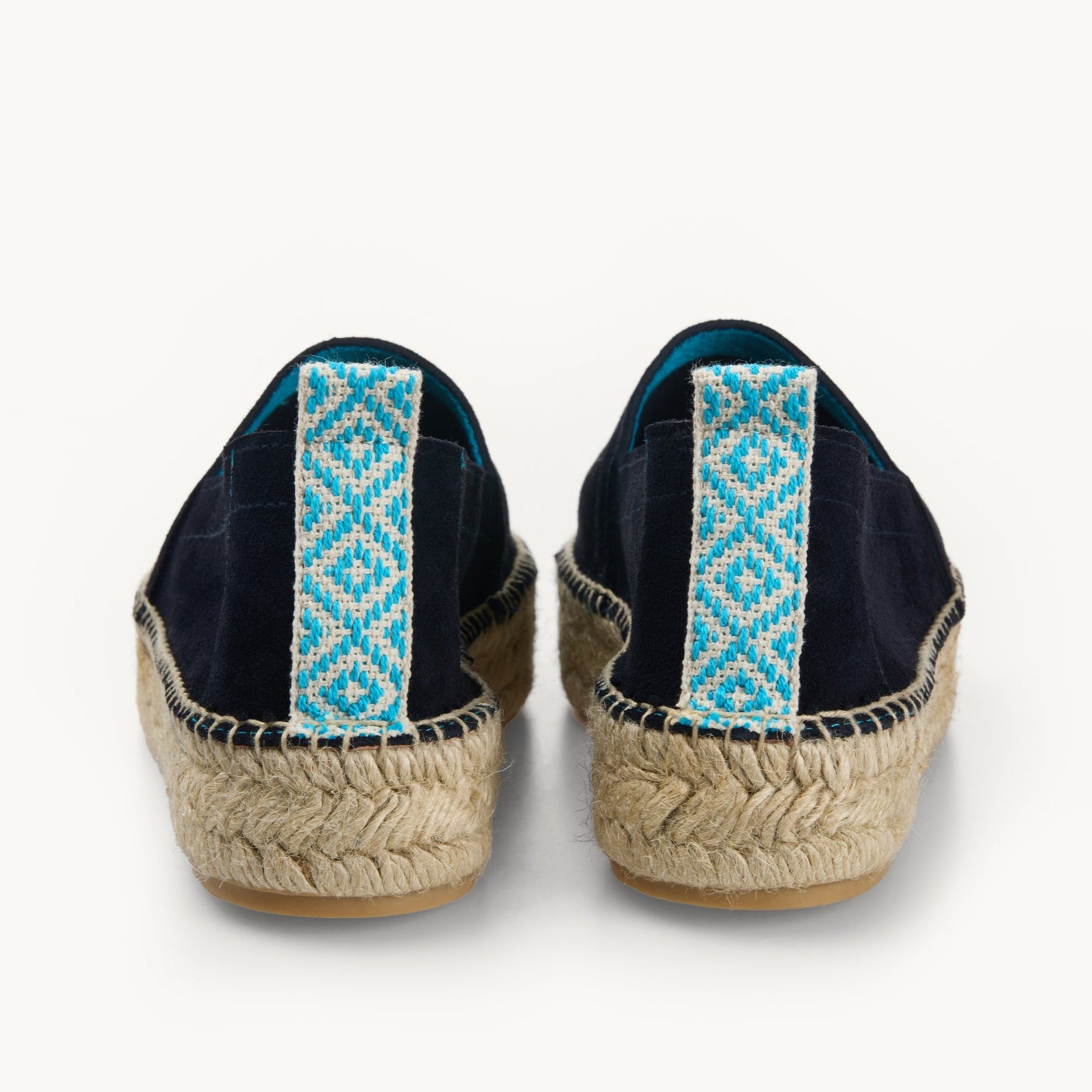 A pair of navy suede women's  espadrille loafers. Only the back details of the heels are shown. A  stripe of turquoise and white patterned ribbon about a centimetre wide runs straight up the back heel. Stitching around the edge of the upper to join it to the  traditional jute sole.  