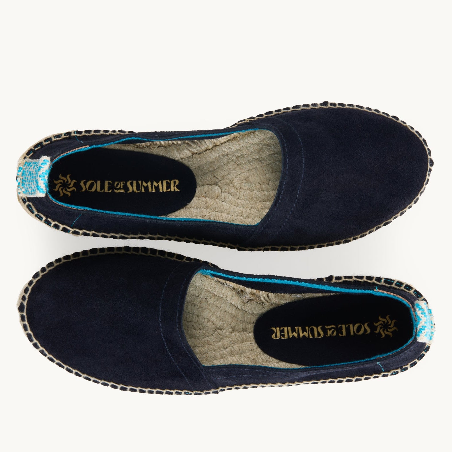 A pair of loafer espadrilles. Flat shoes. Arial view. Navy blue suede upper with turquoise trim around the opening with stitching around the edge joining the upper to the traditional jute sole. Turquoise and white diamond pattern ribbon up the heel of the loafer.