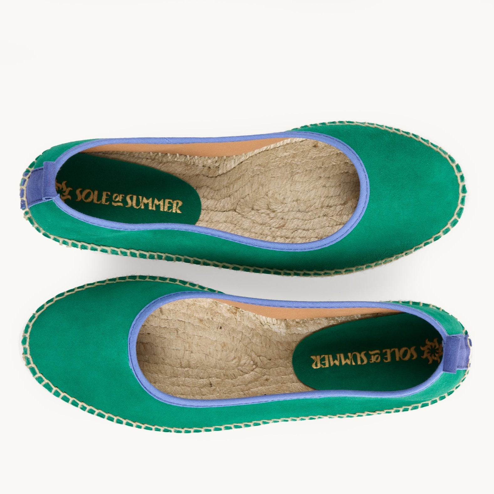 A pair of women's flat ballet espadrilles. View from above looking down on the footwear, unlined jute inside is shown. Green suede upper with blue cotton trim around the opening of the shoe with stitching around the edge joining the upper to the a traditional jute sole. 